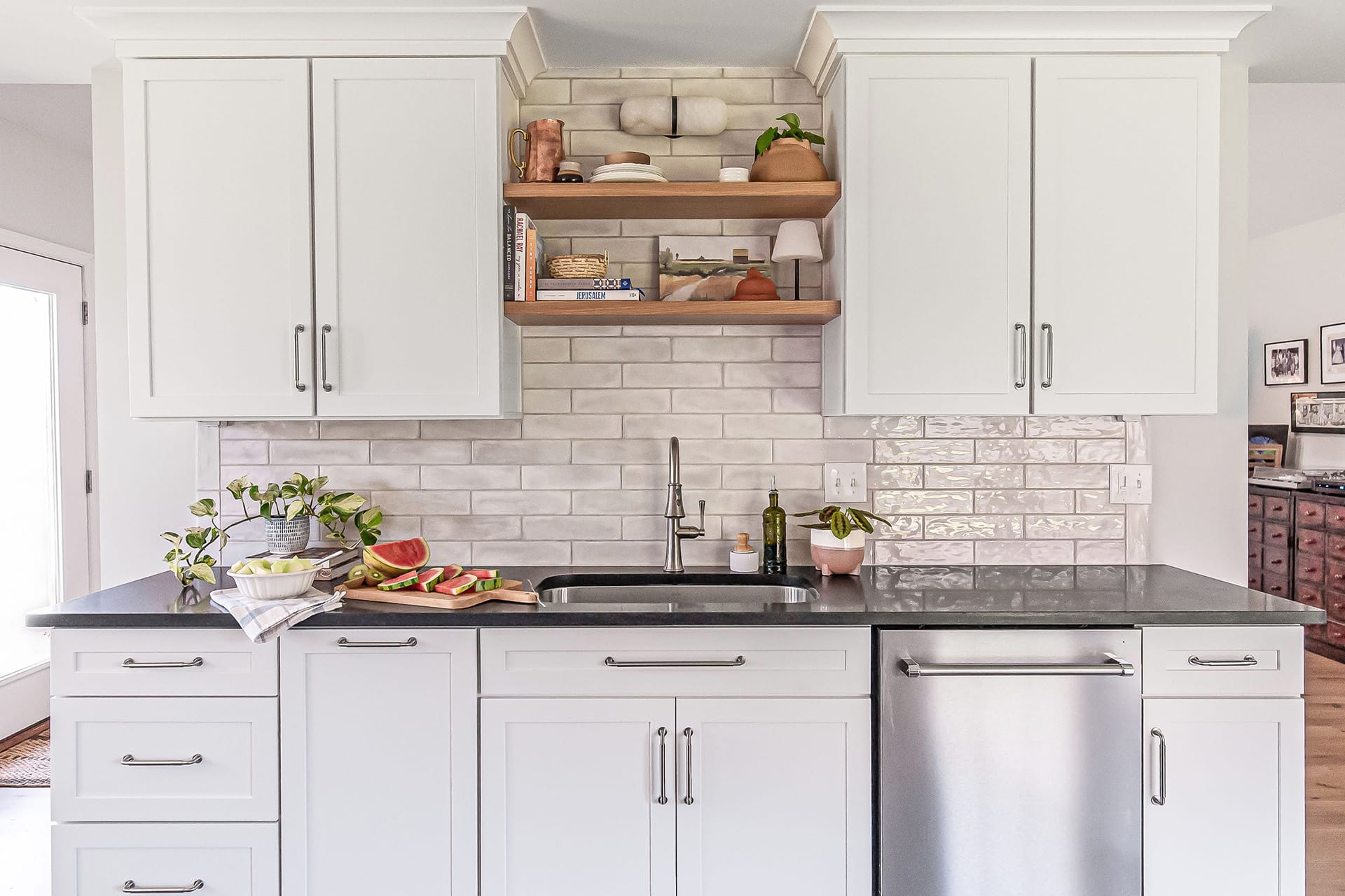 Modern farmhouse kitchen displays bespoke details for everyday function featuring a sink area with open wooden shelves, white cabinetry, and a glossy subway tile backsplash, complimented by fresh watermelon slices on the counter.