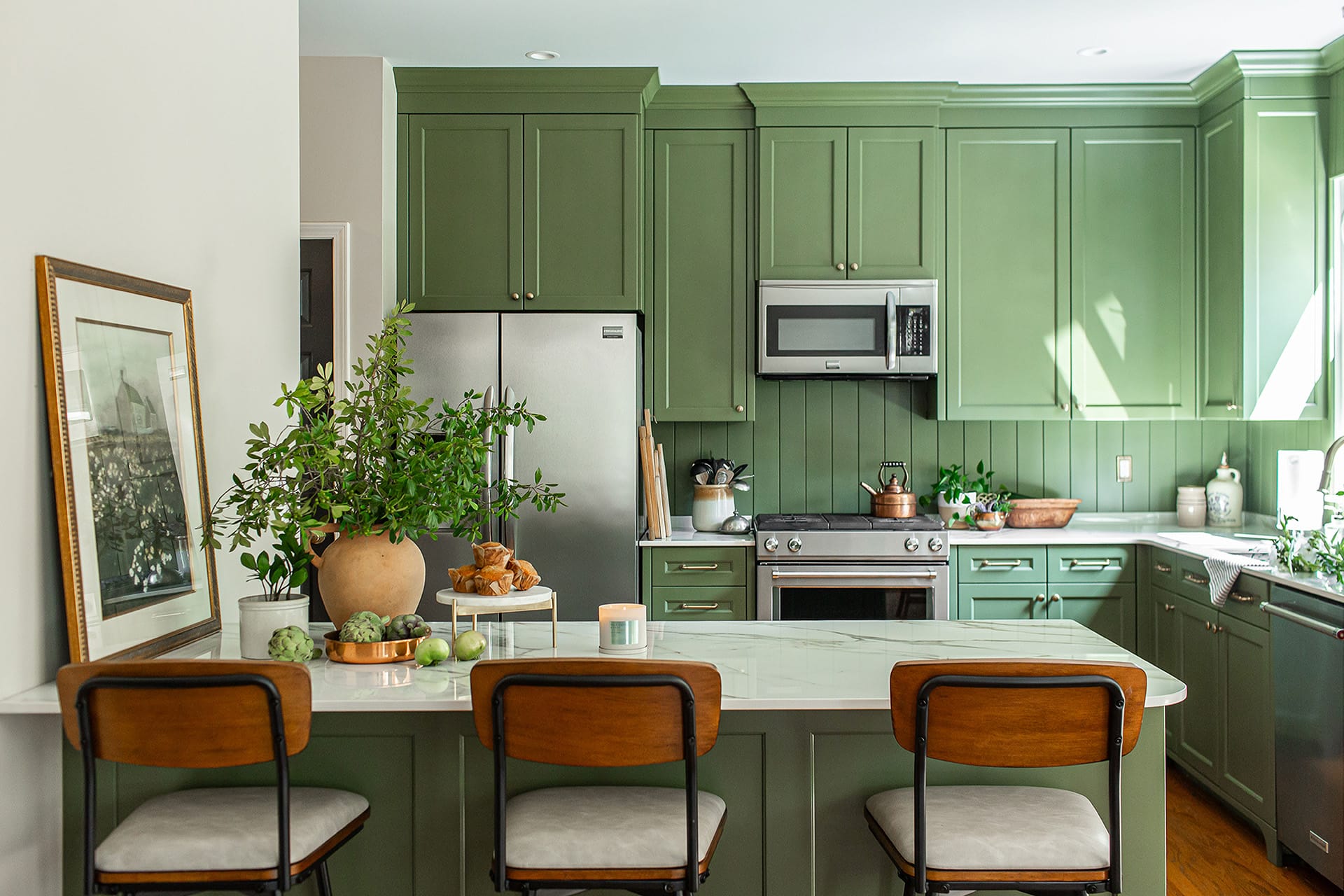 This Brentwood kitchen showcases modern luxury & elegance with green cabinetry, porcelain island, bar seating, and stainless steel appliances featuring stylish natural decor.