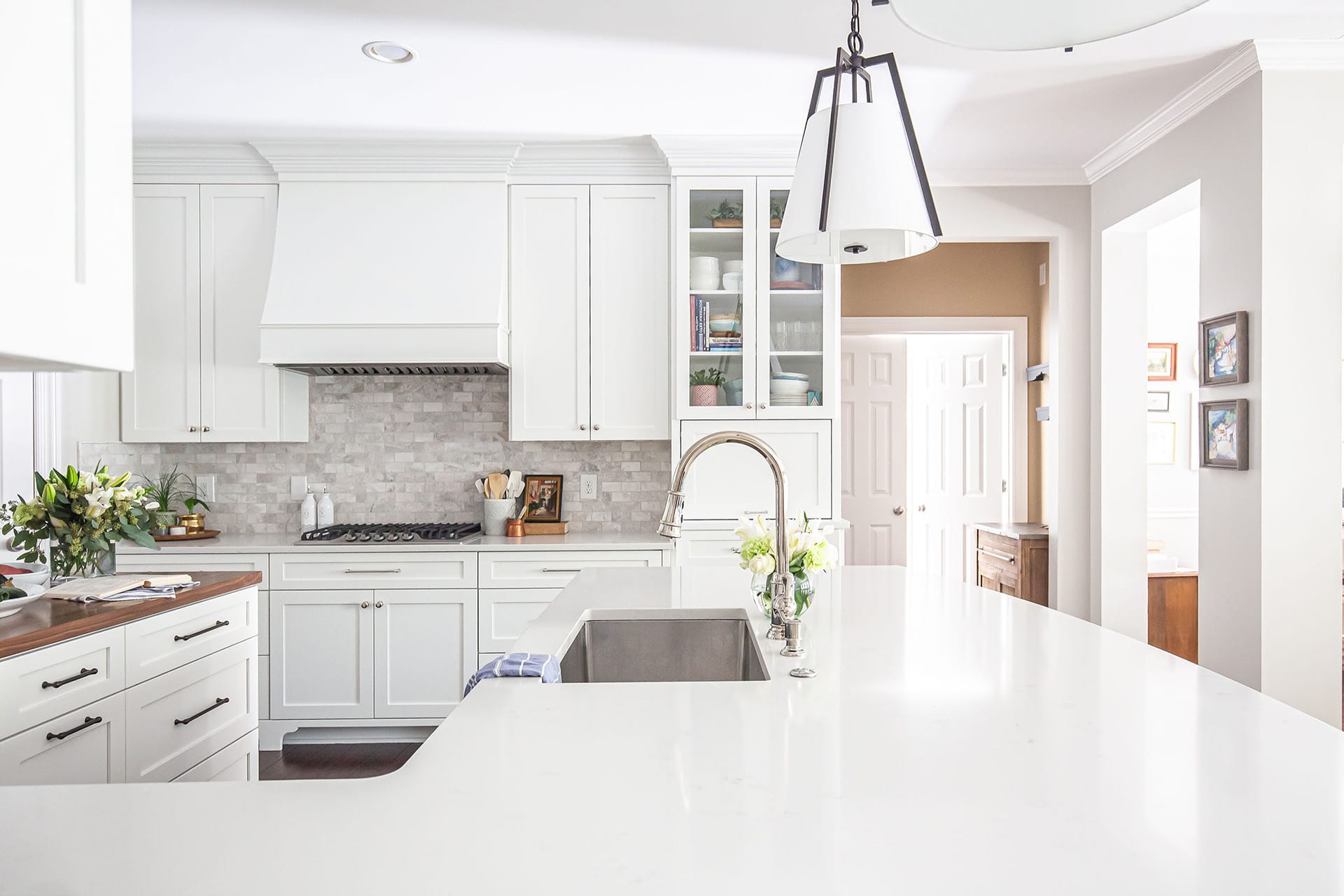 A bright, modern kitchen with white cabinetry, a custom range hood, and marble-style backsplash, complemented by a spacious white quartz island with a stainless steel sink and contemporary faucet.