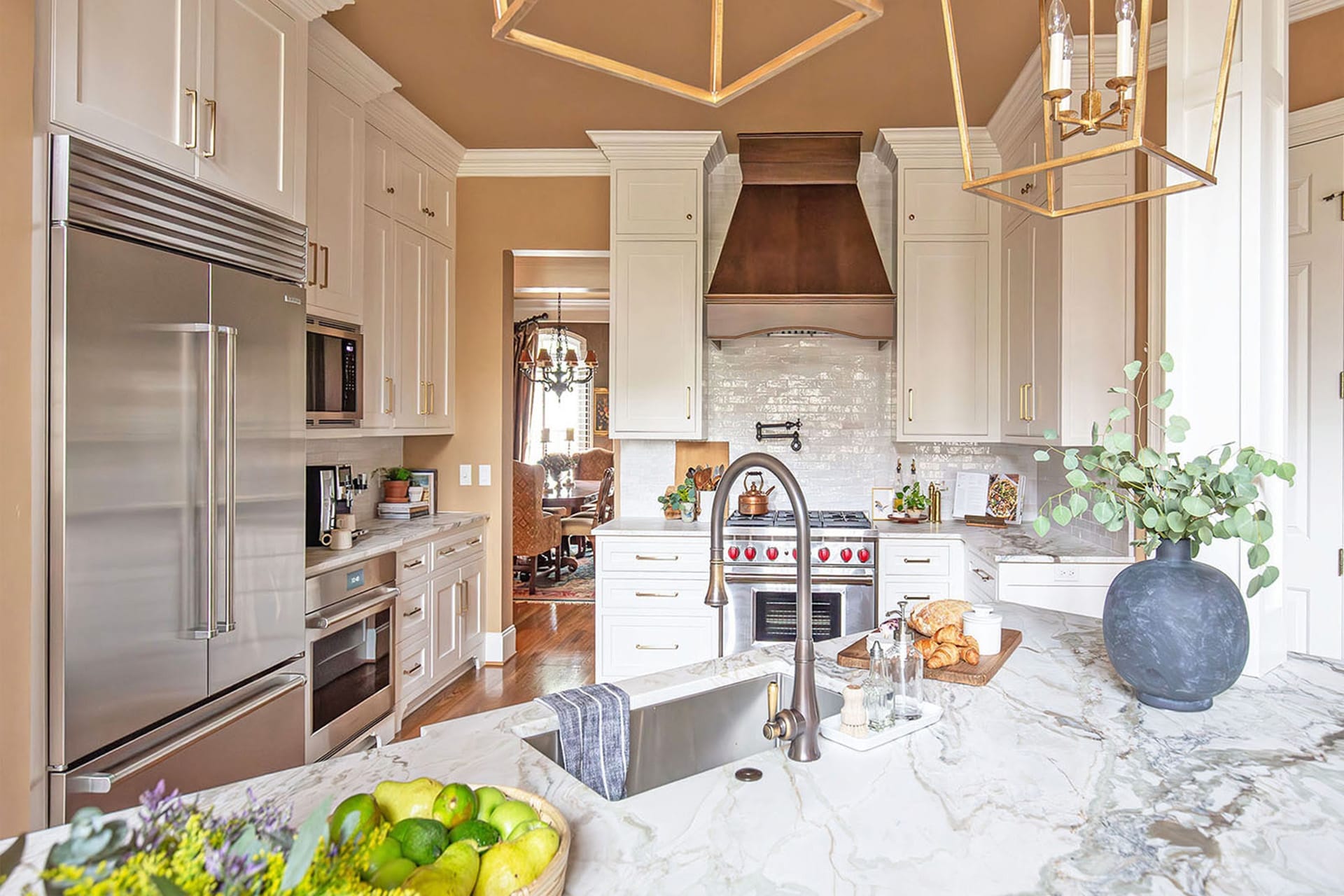 A luxurious kitchen with custom white cabinetry, marble countertops, a wood range hood, and stainless steel appliances, featuring elegant gold accents and natural decor elements.