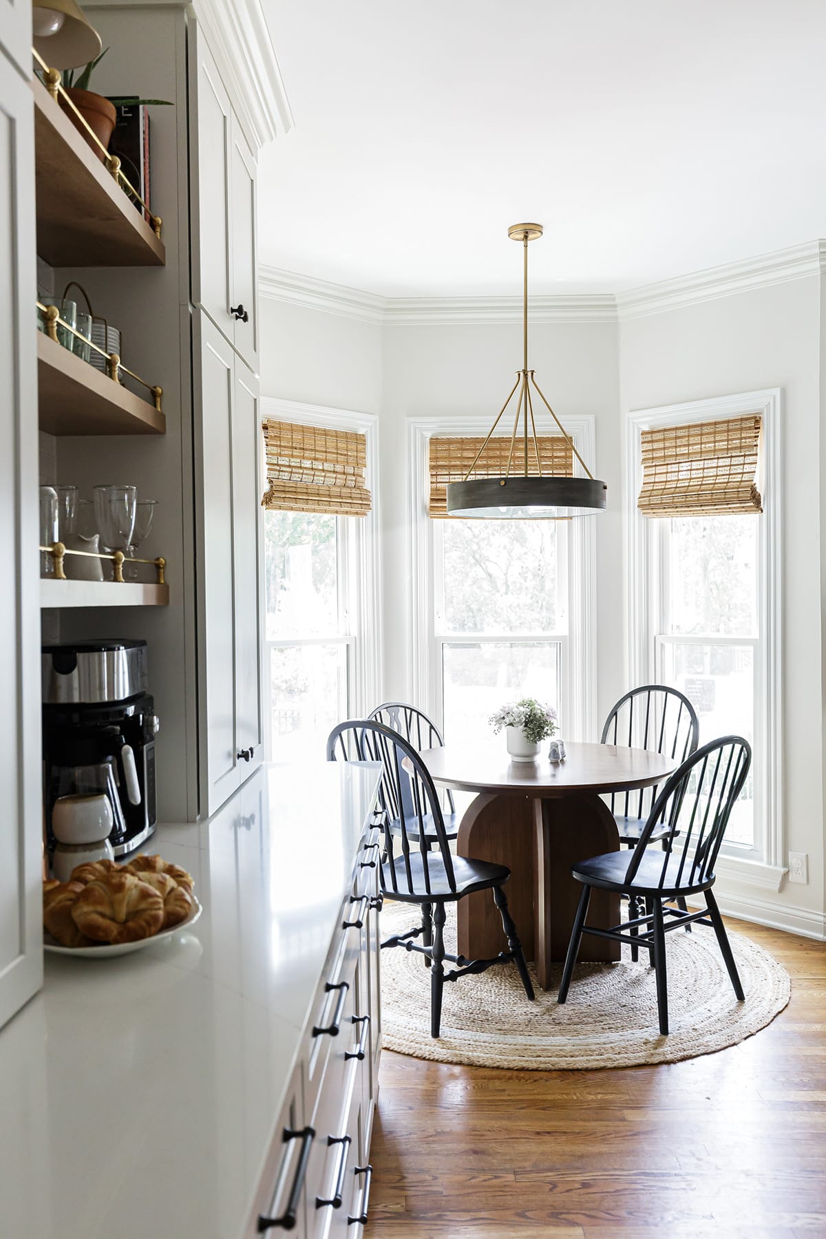 A bright breakfast nook with a round wooden table, black spindle chairs, natural woven shades, and a gold pendant light, adjacent to a kitchen counter with open shelving.