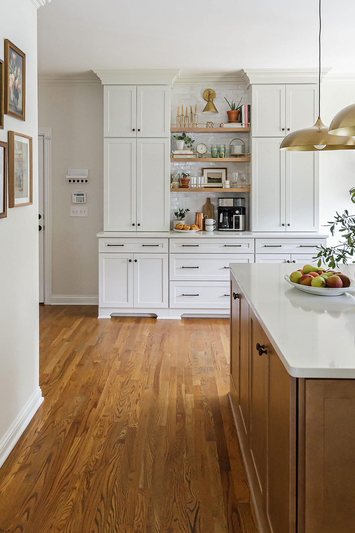 A modern kitchen featuring white cabinetry, a built-in coffee bar with open shelving, brass accents, and a wooden island with a fruit centerpiece.