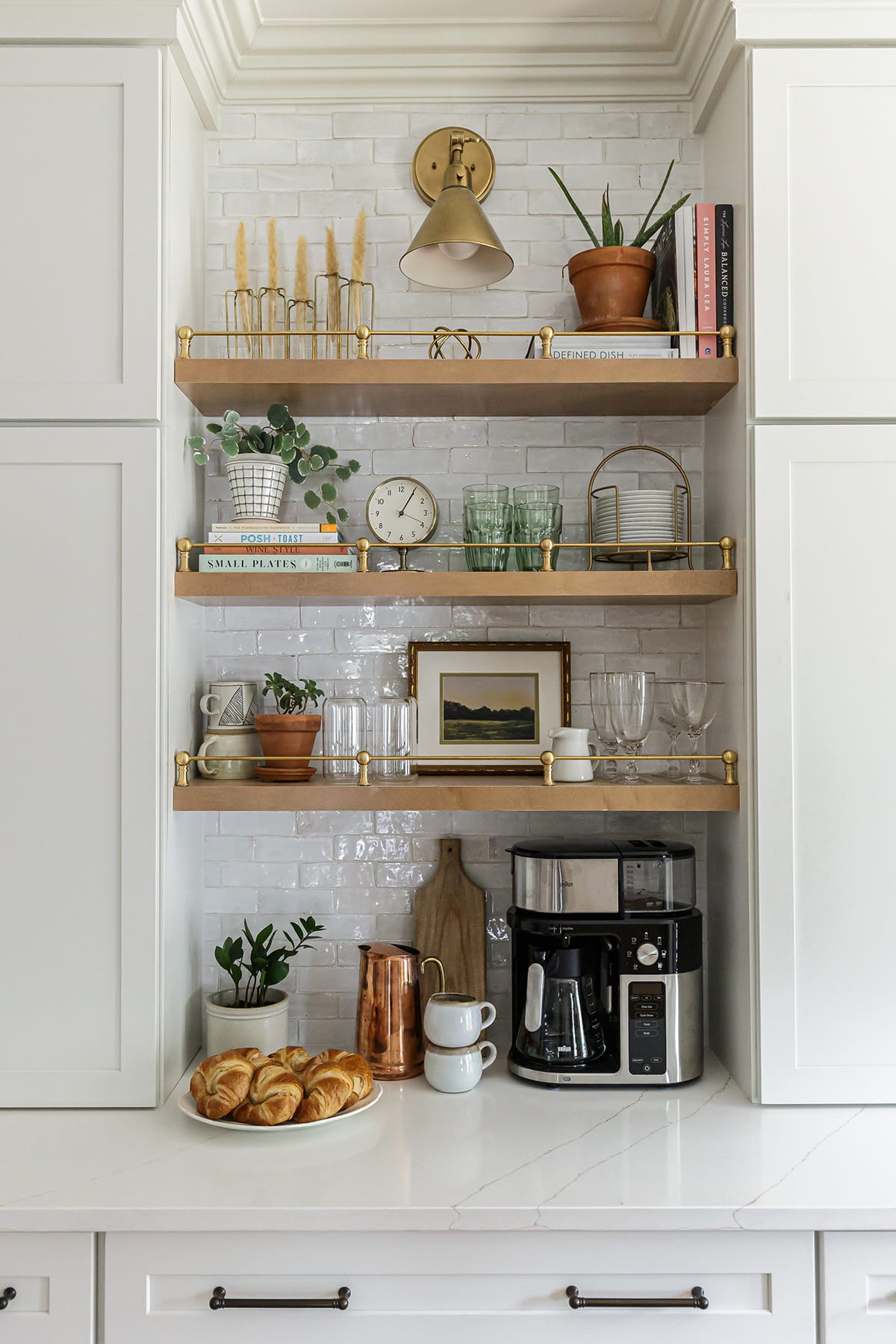 Built-in coffee bar with open wooden shelves featuring brass gallery rails, styled with decor, books, plants, and coffee essentials against a white brick backsplash.