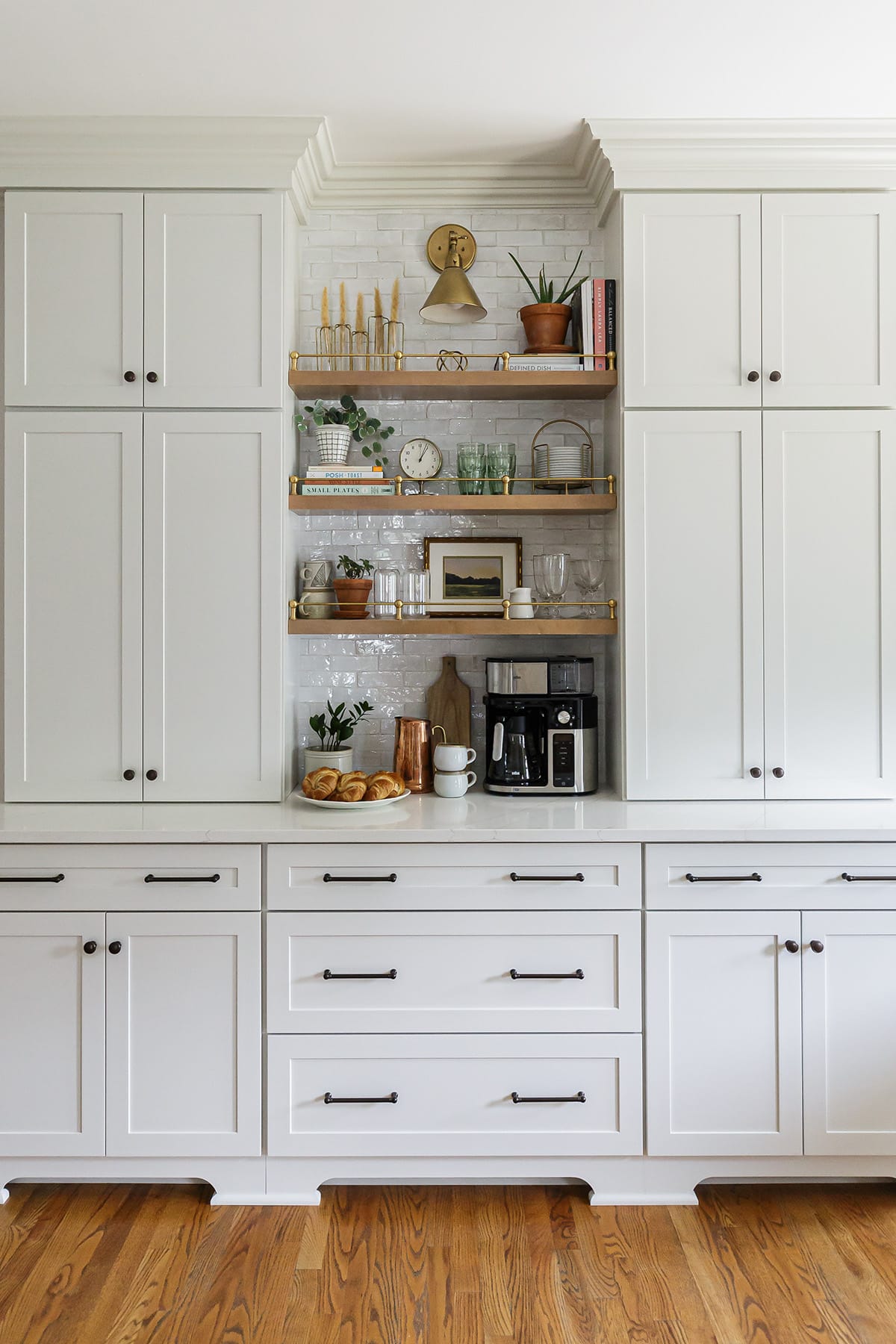 A built-in coffee bar with white cabinetry, open wooden shelves, brass accents, and curated decor, including books, plants, and glassware.