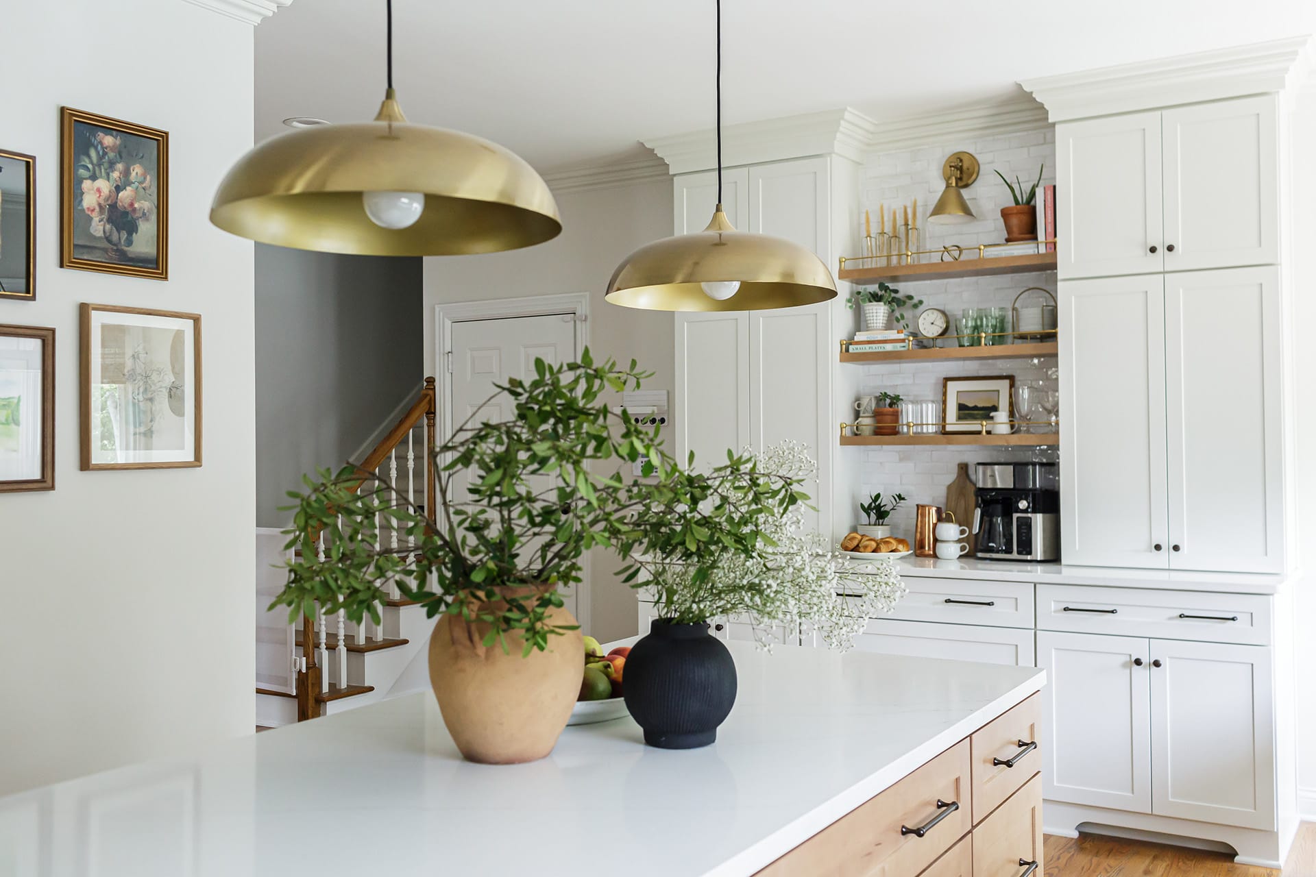 Kitchen view featuring a natural wood island with greenery in decorative vases, brass pendant lights, and a white coffee bar with open shelving.