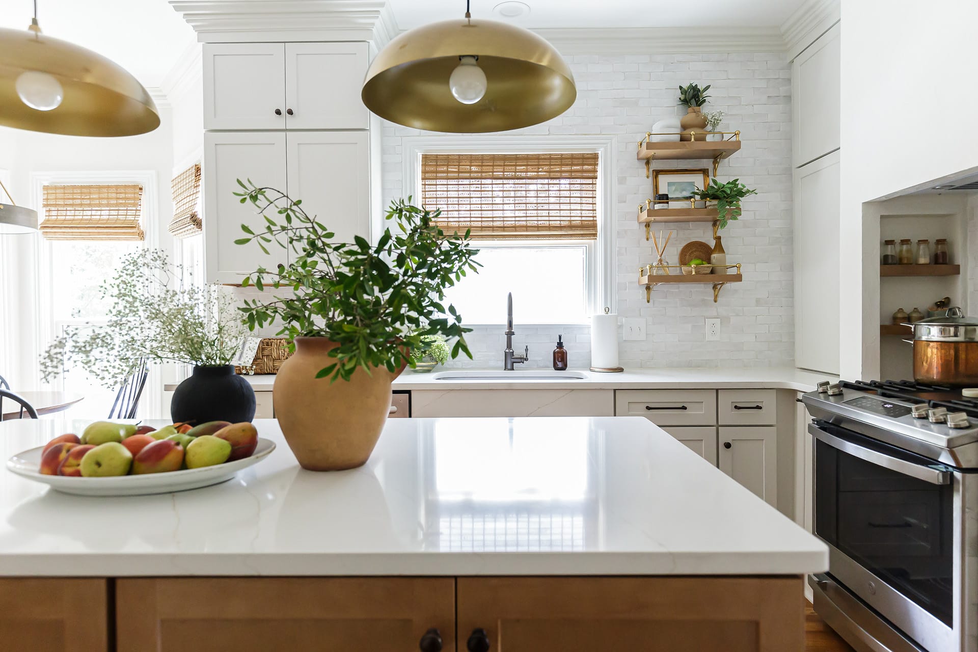 A bright kitchen with a white island, gold pendant lighting, open shelving, and natural accents, including greenery and fresh fruit.