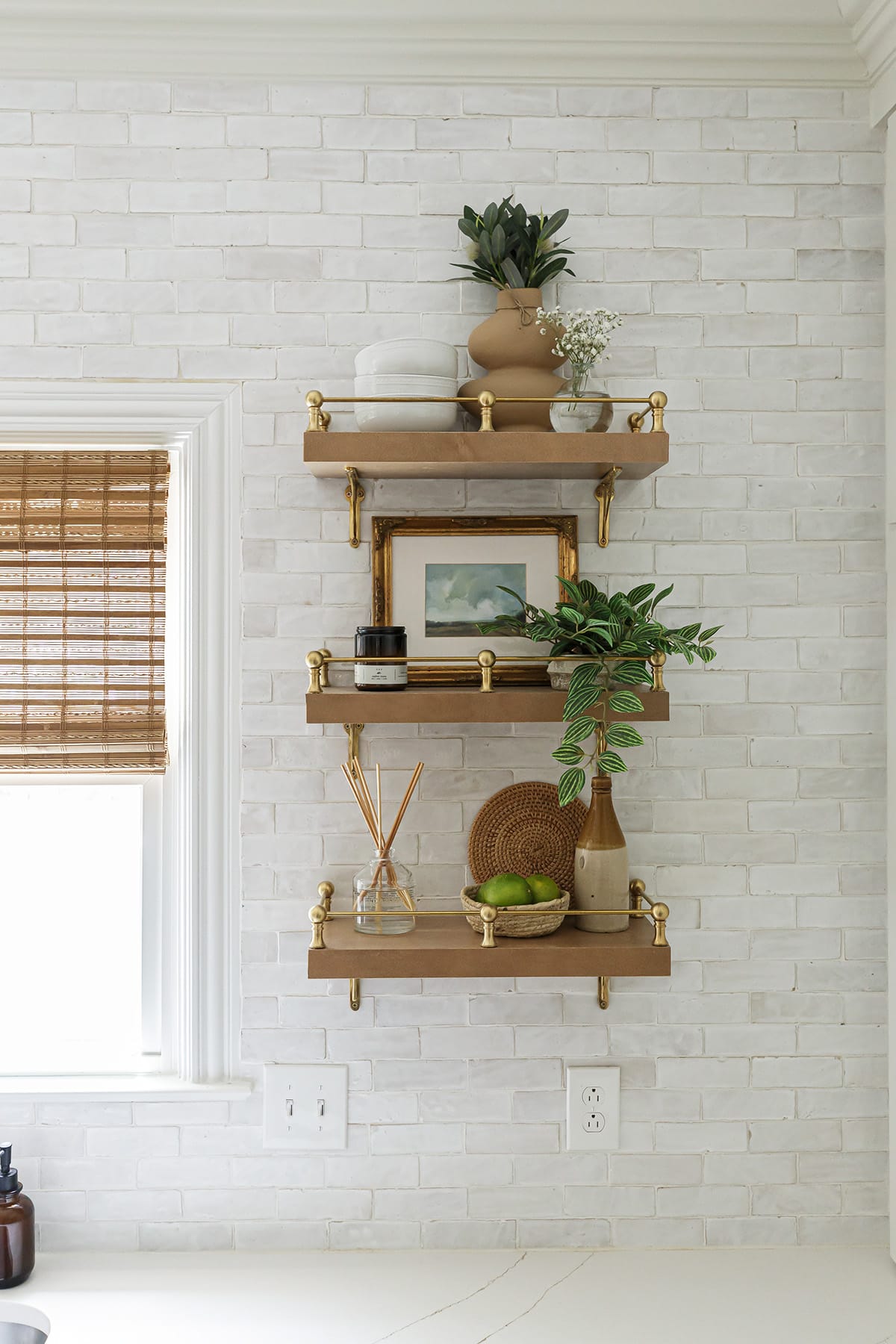 Close-up of open wooden shelves with brass gallery rails, styled with decor items, plants, and artwork against a white brick backsplash.