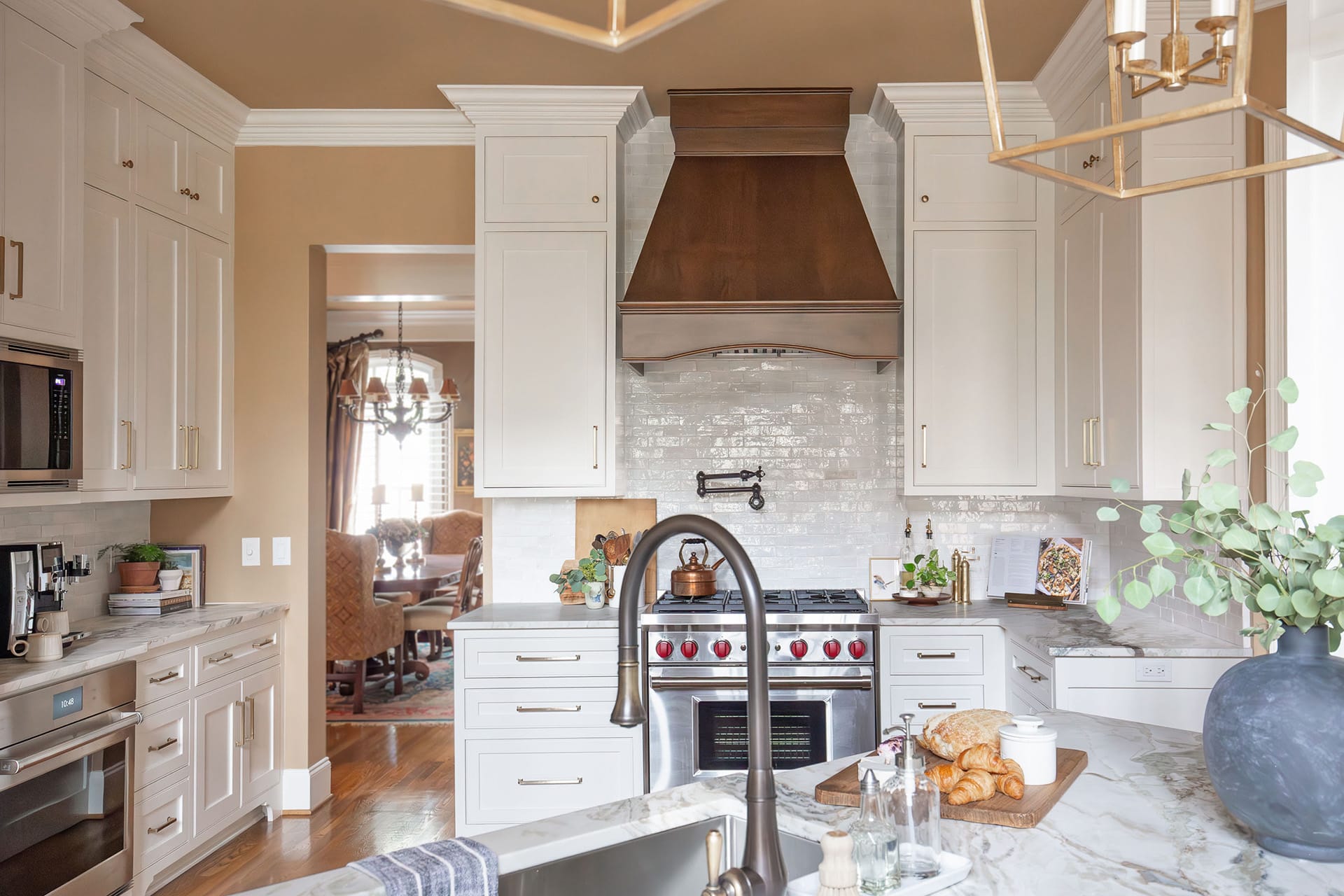 A custom kitchen with white cabinetry, a rich wood range hood, marble countertops, and stainless steel appliances, styled with gold accents and natural decor elements.