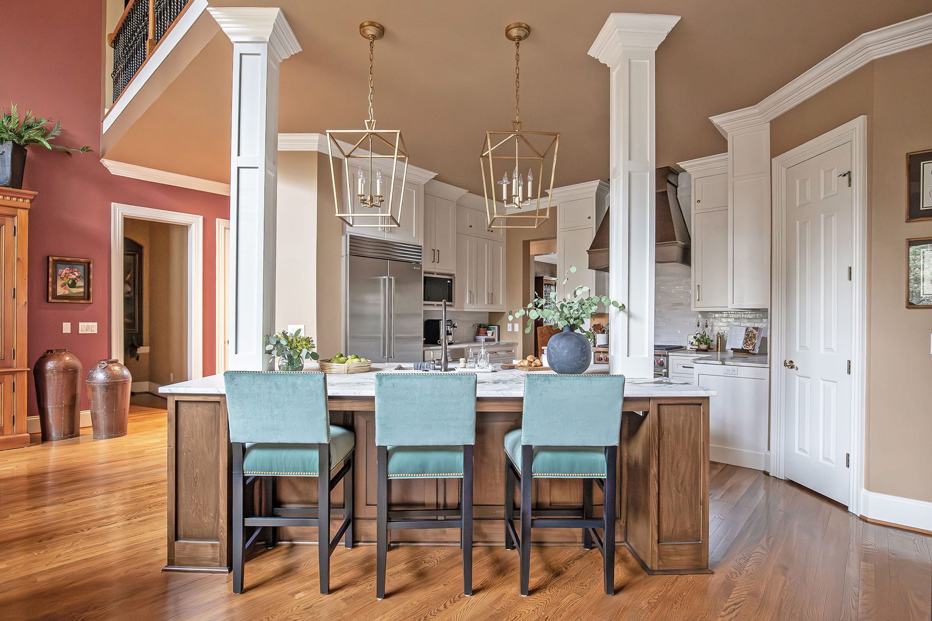 A spacious kitchen with a large wood island, marble countertop, teal upholstered bar stools, gold pendant lighting, and custom white cabinetry.