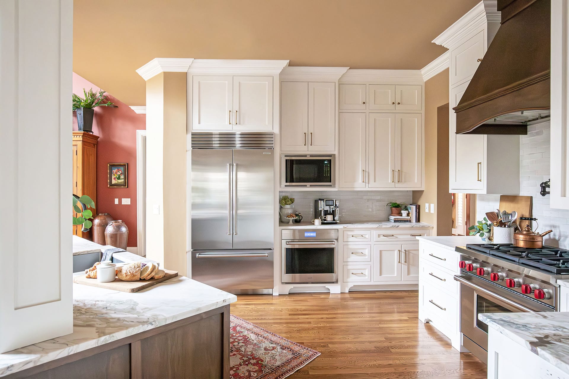 A spacious kitchen featuring white cabinetry, stainless steel appliances, a bronze range hood, marble countertops, and warm wood flooring, styled with natural accents.