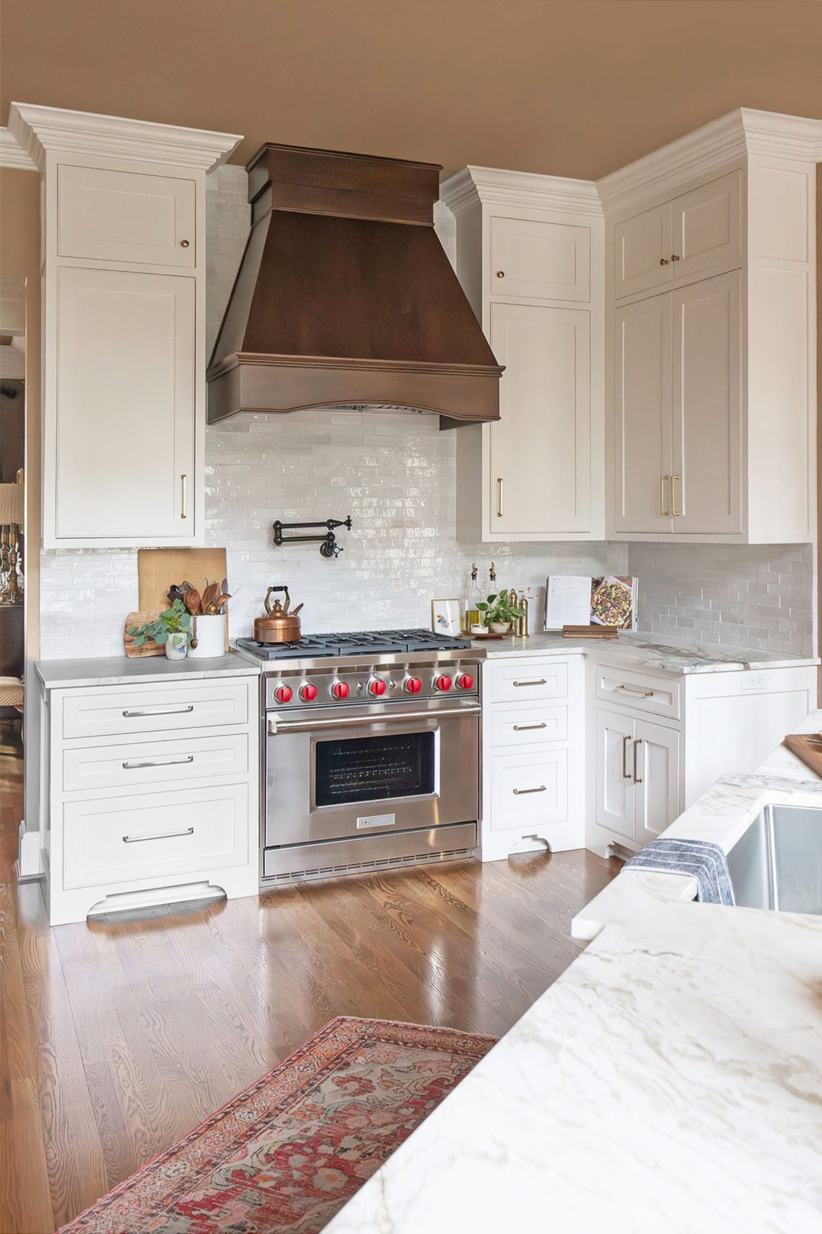 A luxurious kitchen featuring a wood range hood, white cabinetry, stainless steel Wolf range, marble countertops, and warm wood flooring.