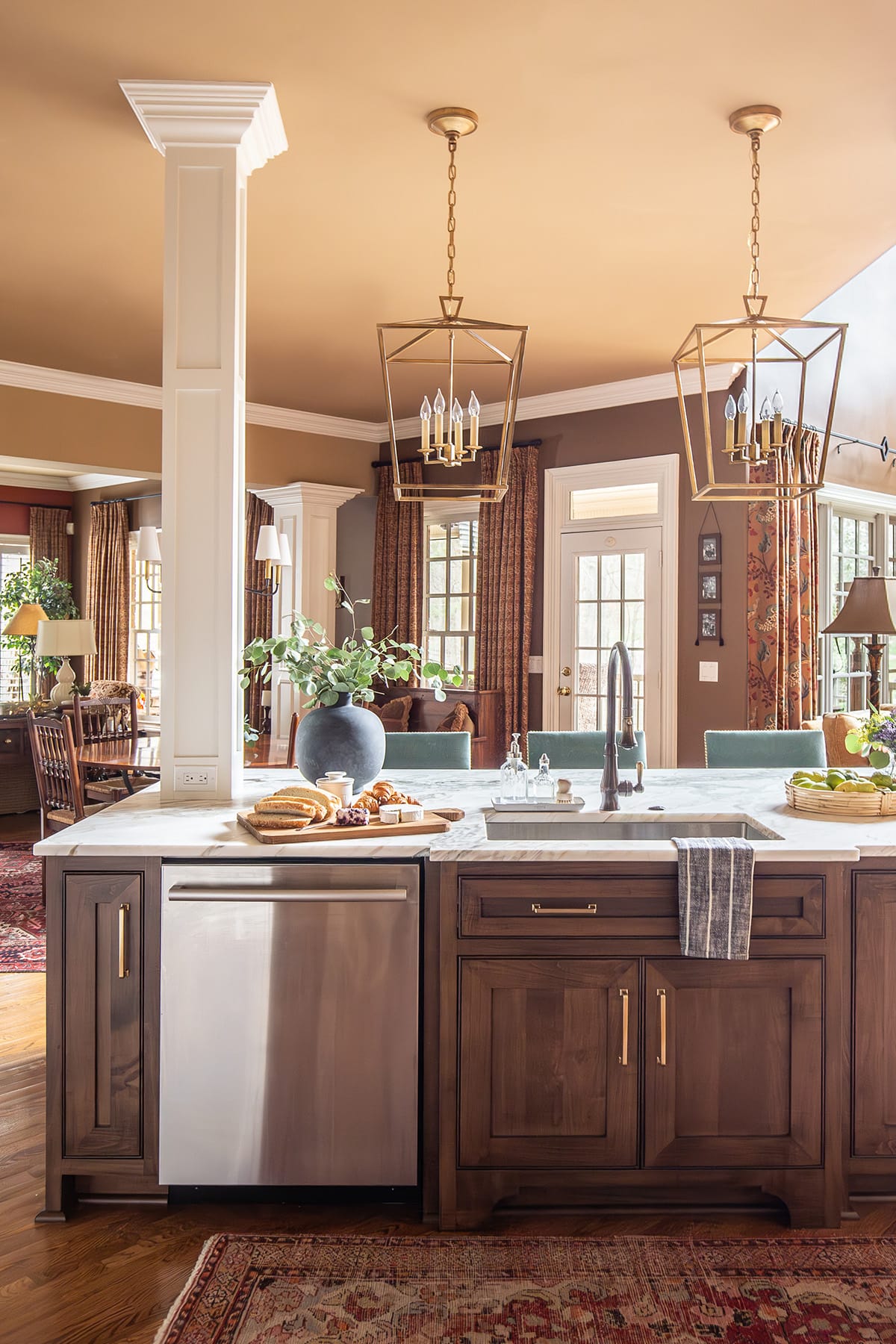 A custom kitchen island with rich wood cabinetry, marble countertops, a stainless steel dishwasher, bronze faucet, and gold pendant lighting, set against a warm, inviting dining and living area.