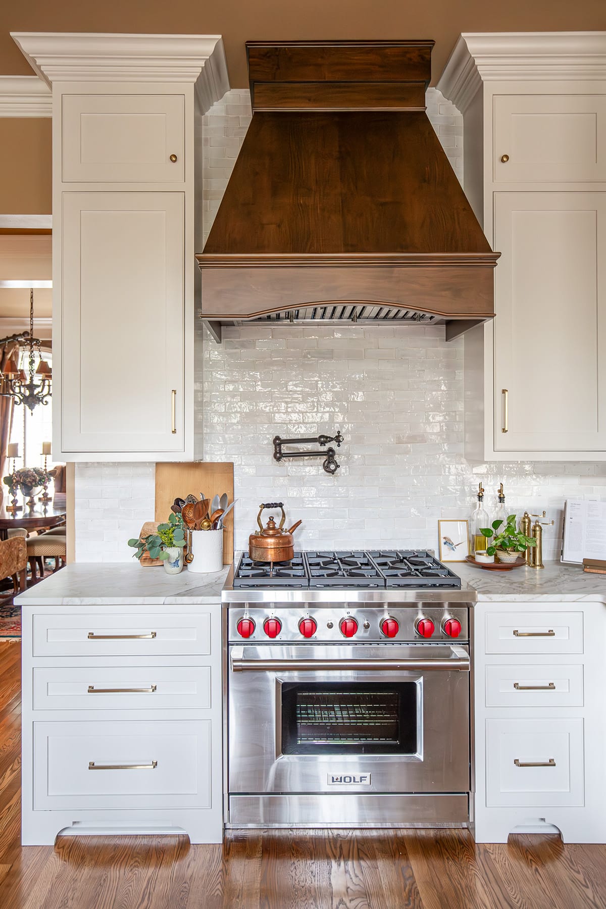 A custom kitchen that exudes timeless style for inspired living with a rich wood range hood, white cabinetry, a professional-grade Wolf range, and a glossy white tile backsplash, styled with copper and brass accents.