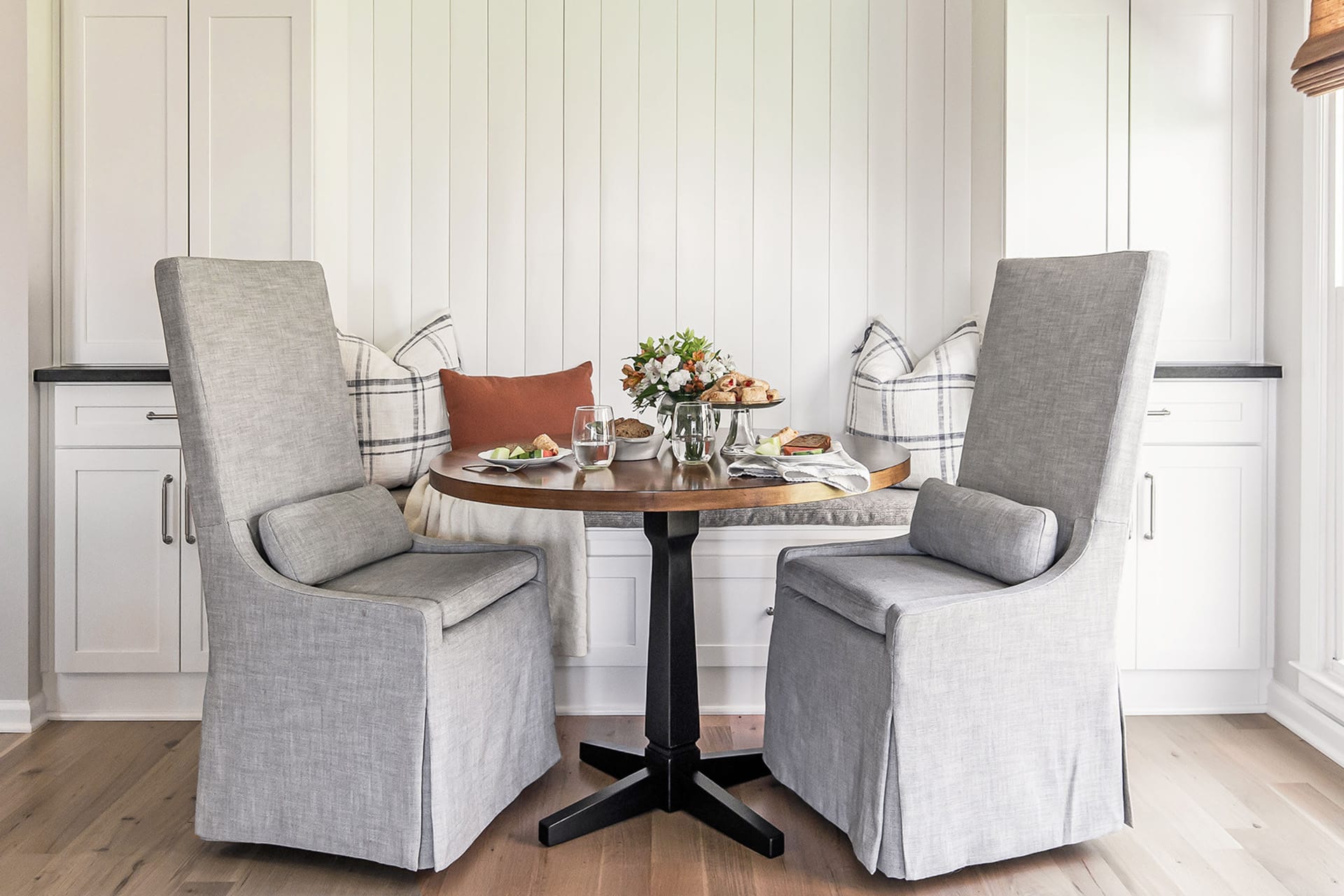 Cozy farmhouse breakfast nook with a round wooden table, cushioned bench seating, and upholstered high-back chairs, styled with plaid pillows and fresh flowers.