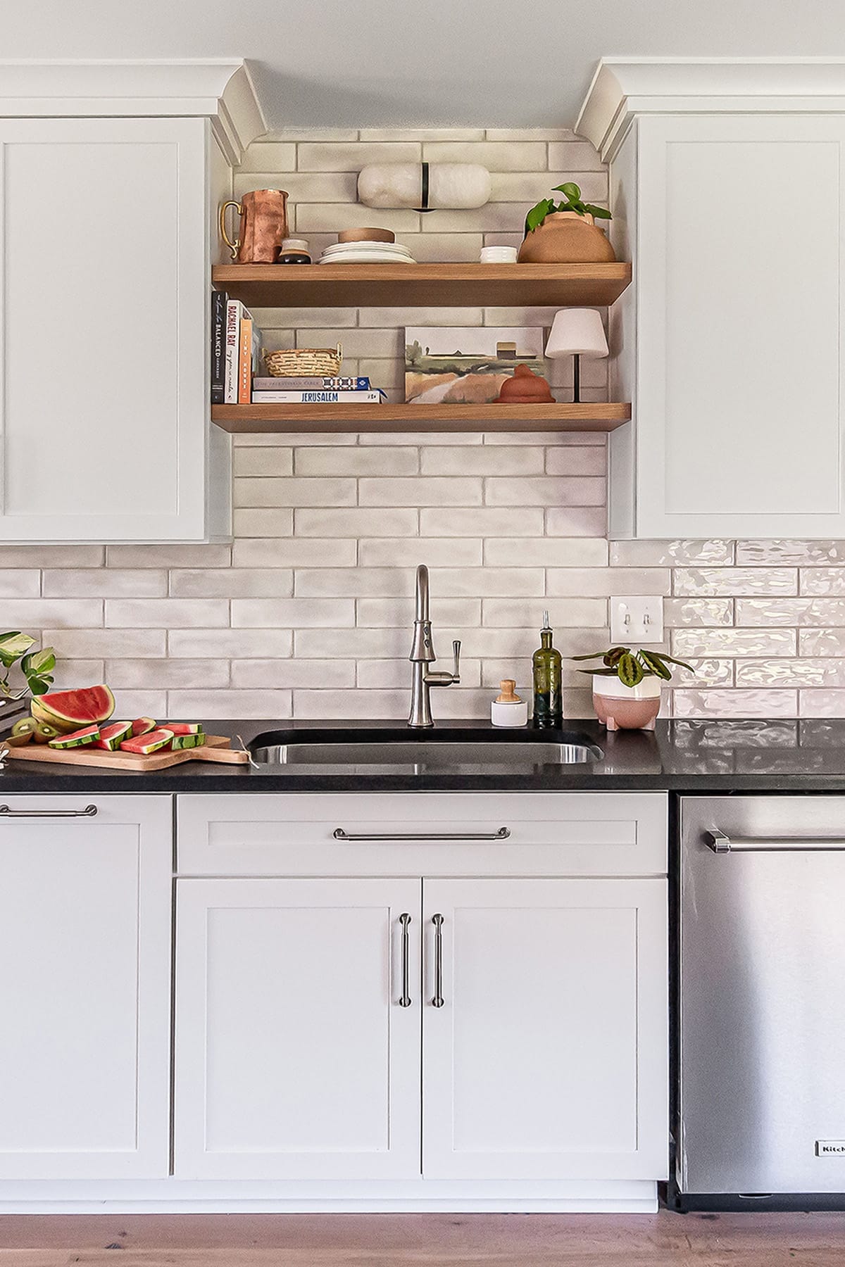 A modern farmhouse kitchen with white cabinetry, a dark countertop, and open wooden shelves styled with natural accents, books, and ceramics, set against a handcrafted subway tile backsplash.