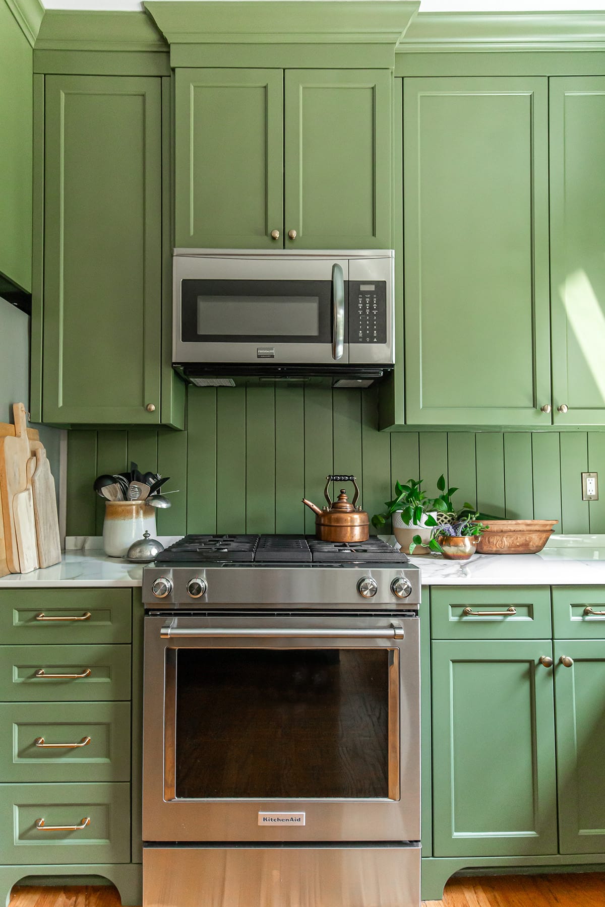 Close-up of a green kitchen with stainless steel range, microwave, porcelain countertops, and natural decor accents.