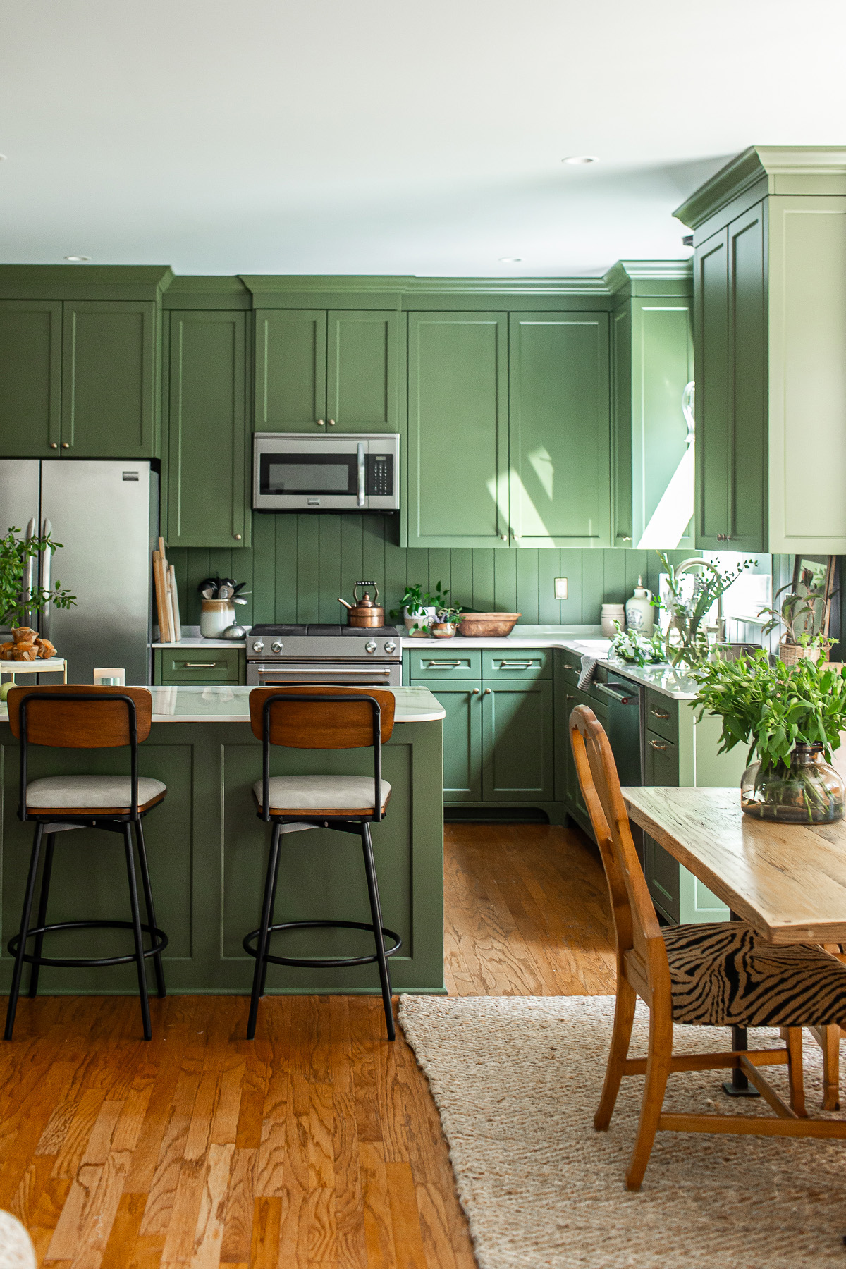 Full view of a green kitchen with bar seating, stainless steel appliances, porcelain countertops, and natural wood and greenery accents.