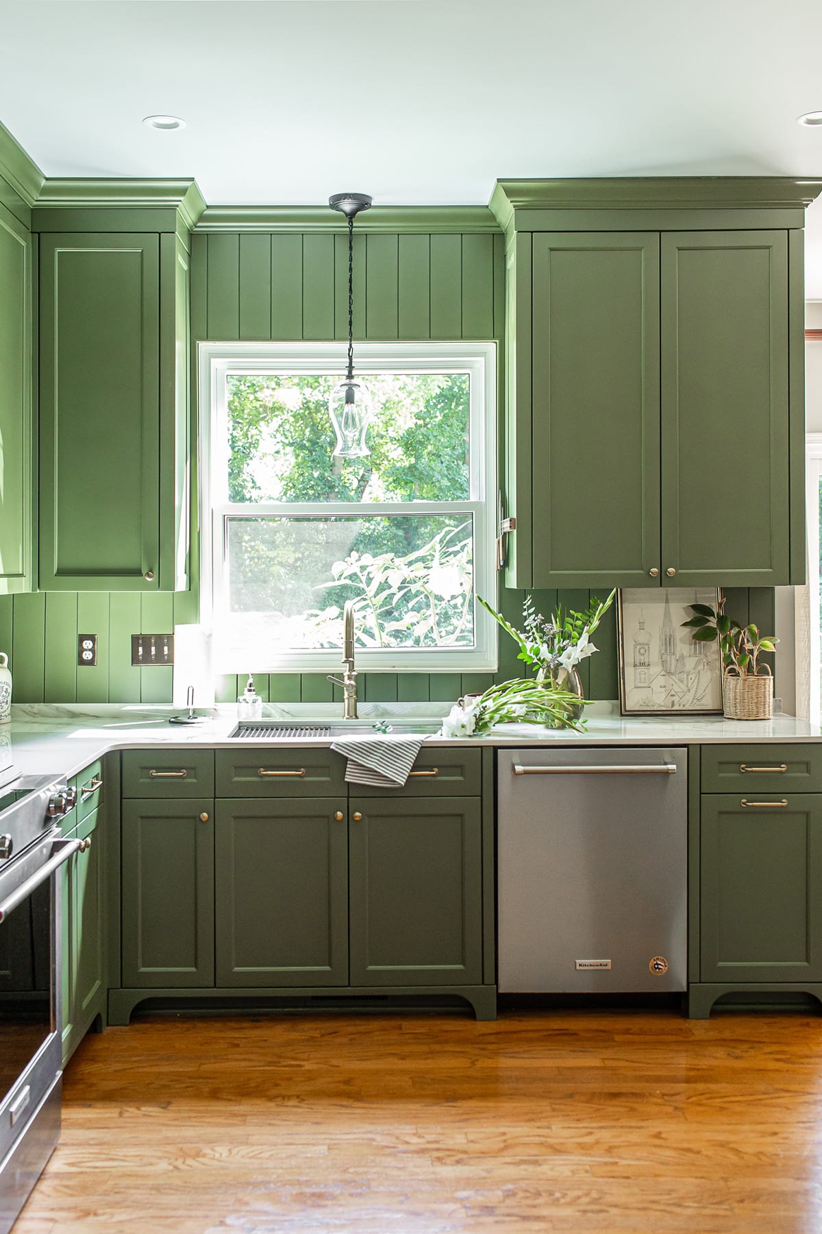 Green kitchen with a large window, stainless steel dishwasher, porcelain countertops, and natural light highlighting fresh greenery.