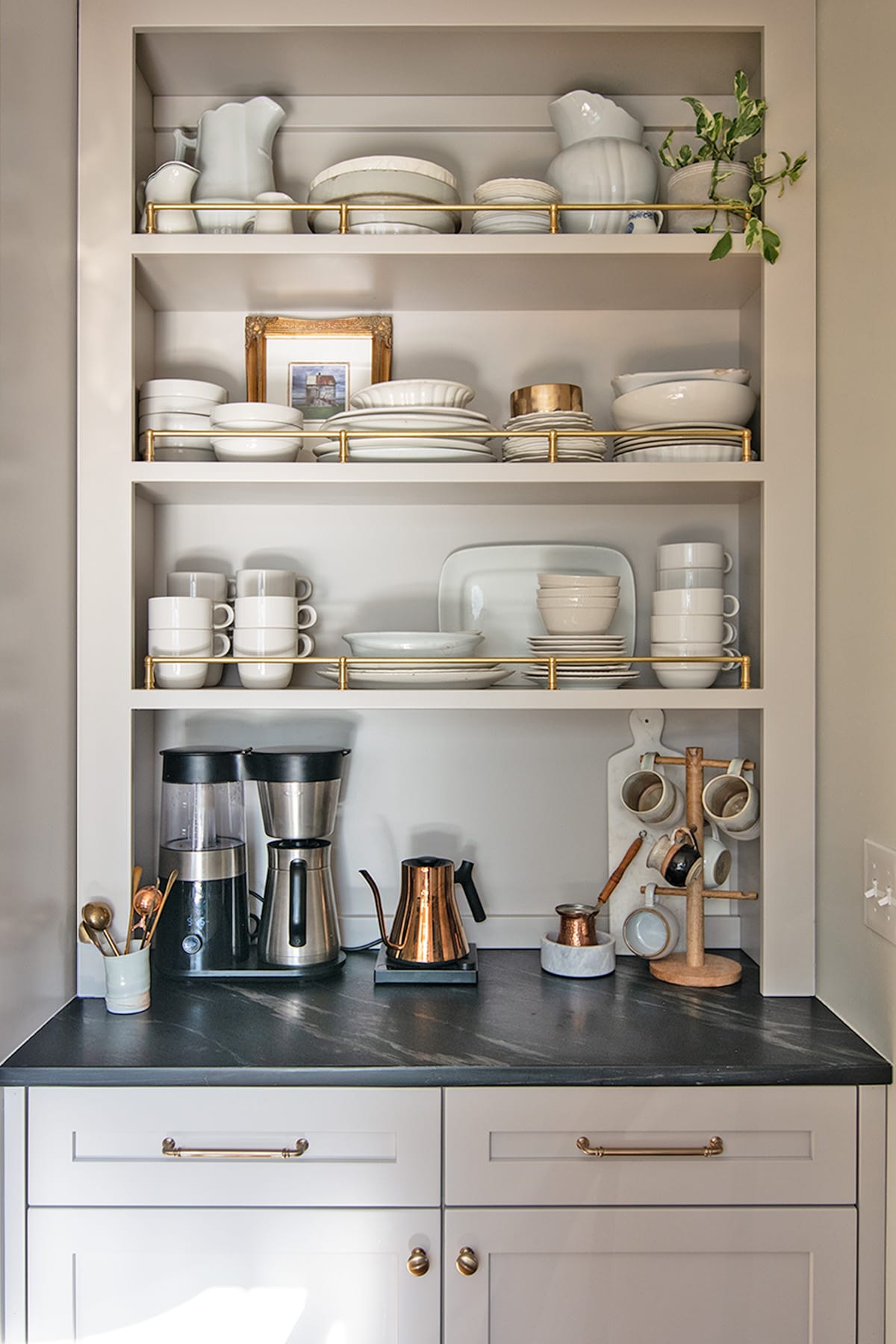 A beautifully organized coffee bar featuring light gray cabinetry, ironstone dishes displayed on open shelving with brass accents, and coffee essentials on a dark countertop.