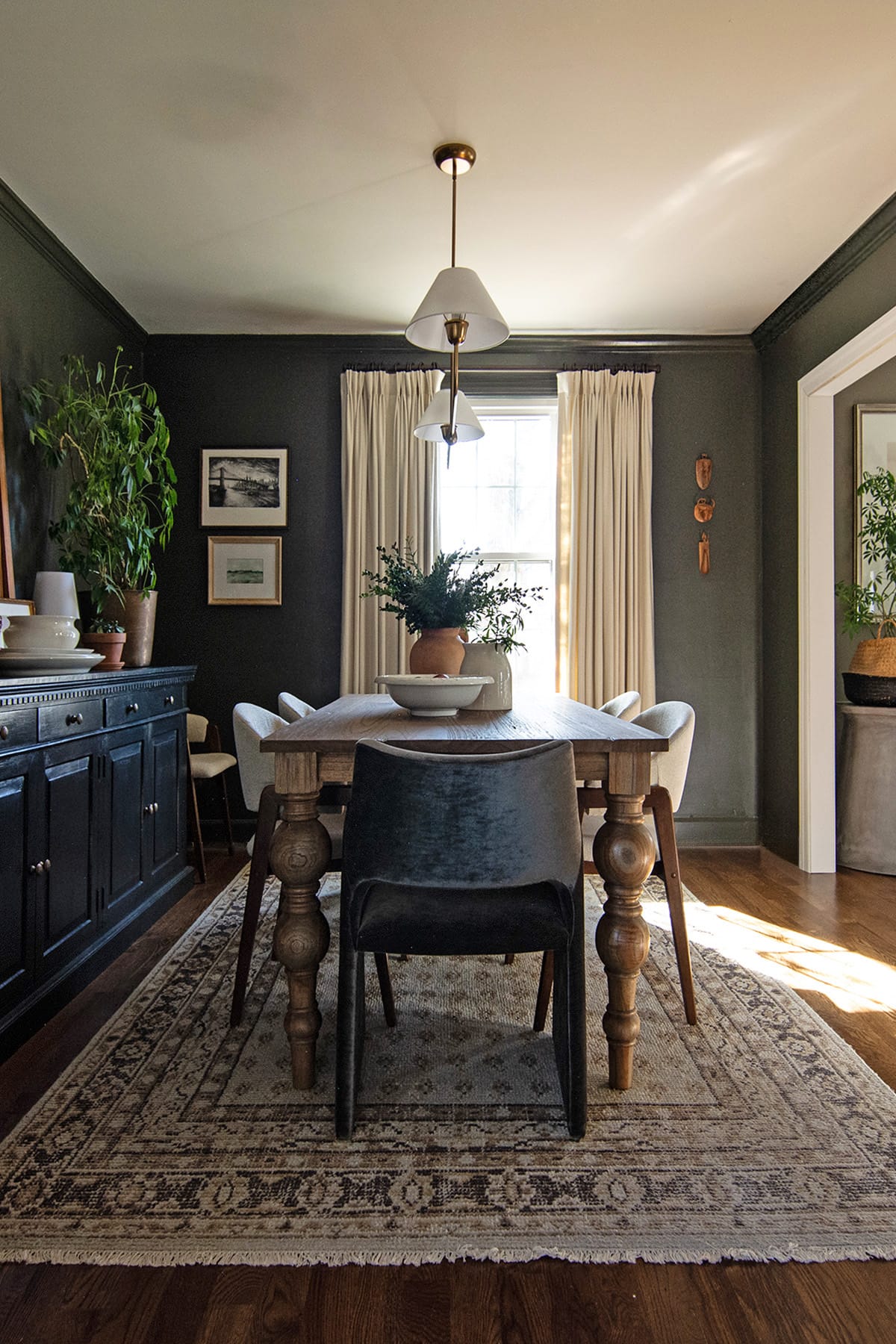 A dark green dining room with a wooden table featuring turned legs, velvet and upholstered chairs, and a window framed by cream curtains that bring in natural light.