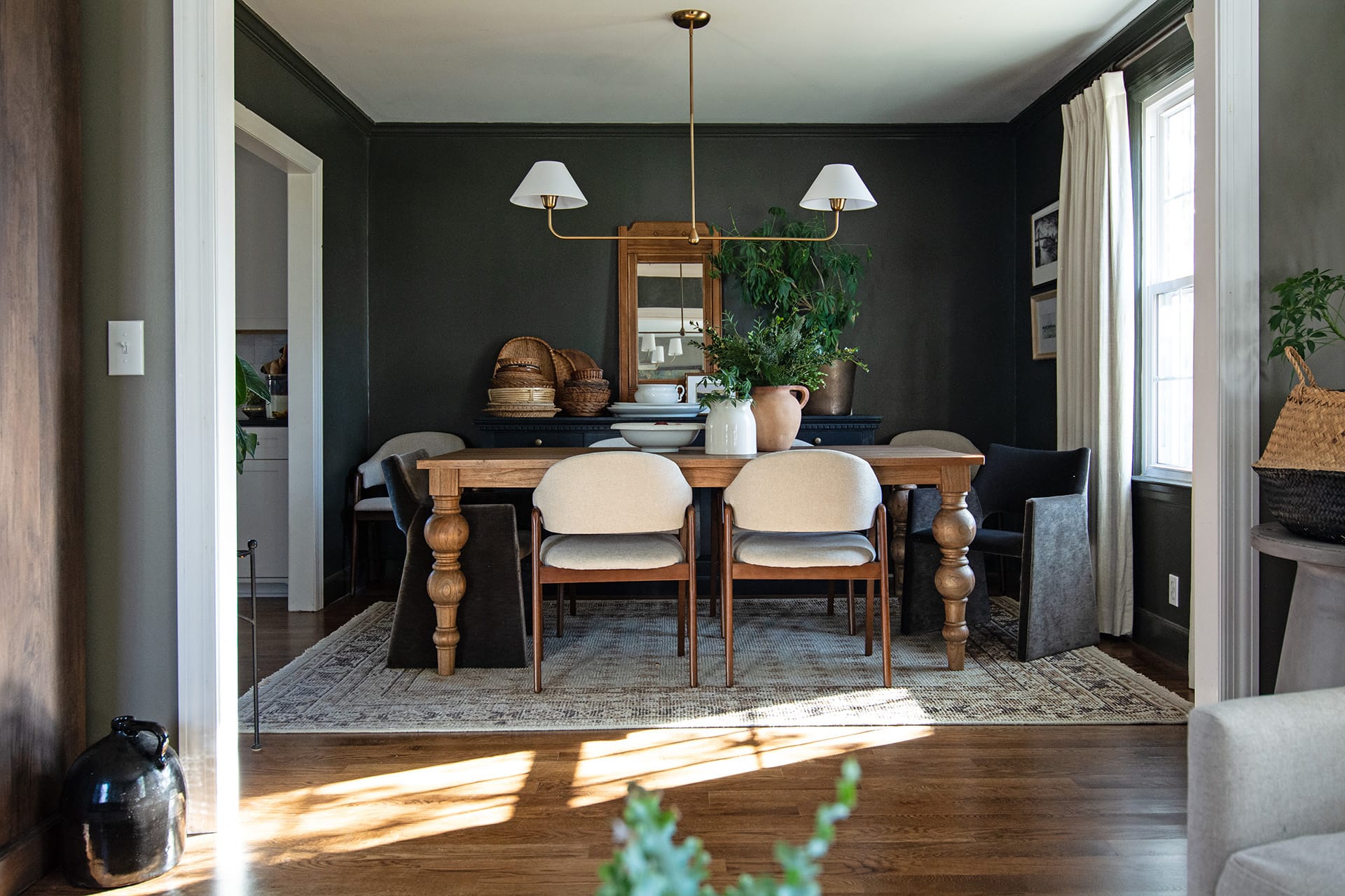 A dark green dining room featuring a wooden table with turned legs, upholstered and velvet chairs, a black buffet with greenery and decor, and a modern brass chandelier.