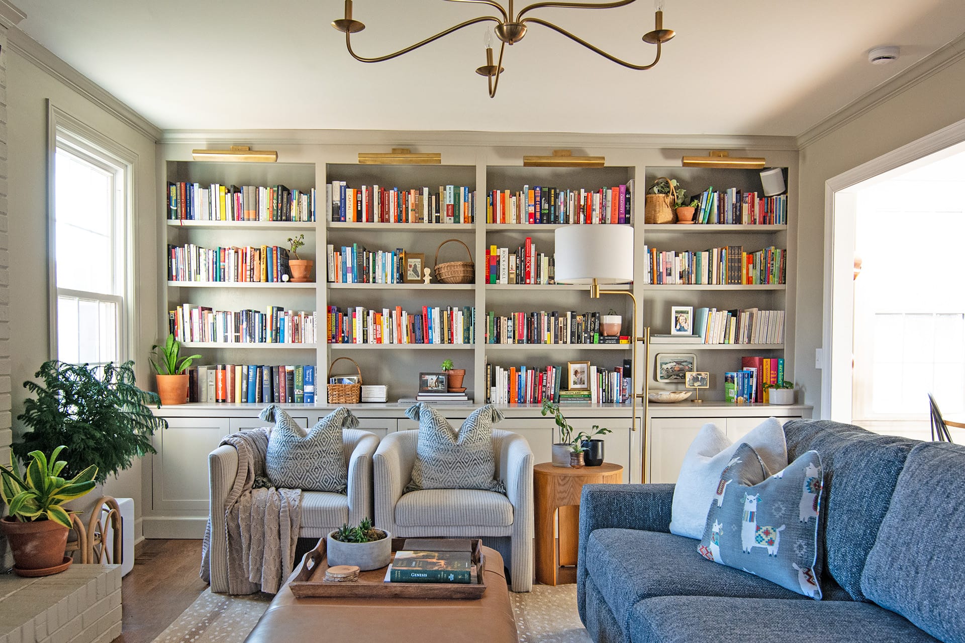 A cozy living room with built-in bookshelves filled with colorful books, armchairs with patterned pillows, a blue sectional sofa, and vibrant green plants.