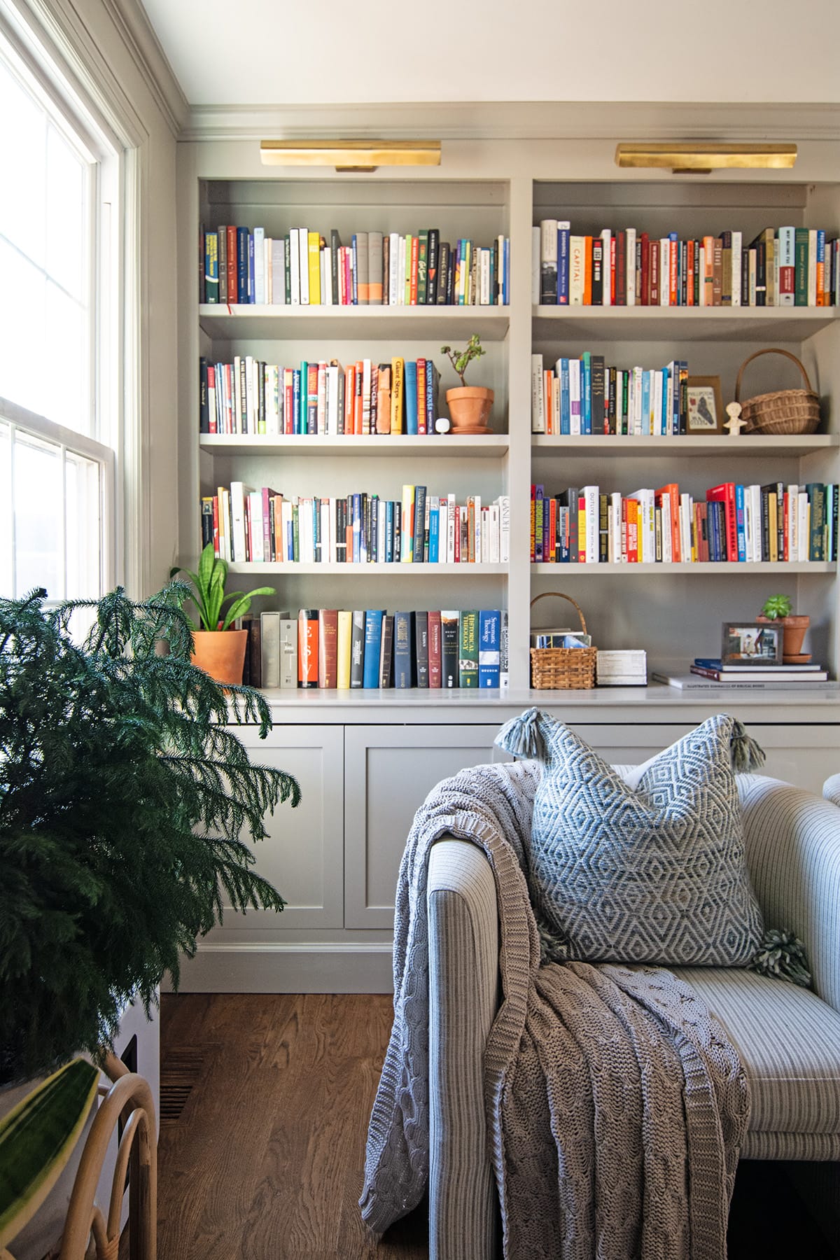 A cozy corner in a living room featuring built-in bookshelves with colorful books, a striped armchair with a patterned pillow and knitted throw, and vibrant greenery.