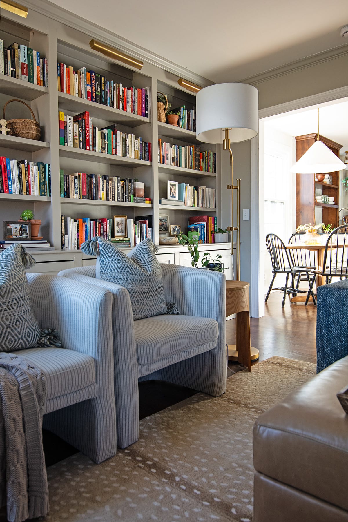 Two light blue striped armchairs with patterned pillows in front of built-in bookshelves filled with colorful books, a gold floor lamp, and a glimpse into the dining area.