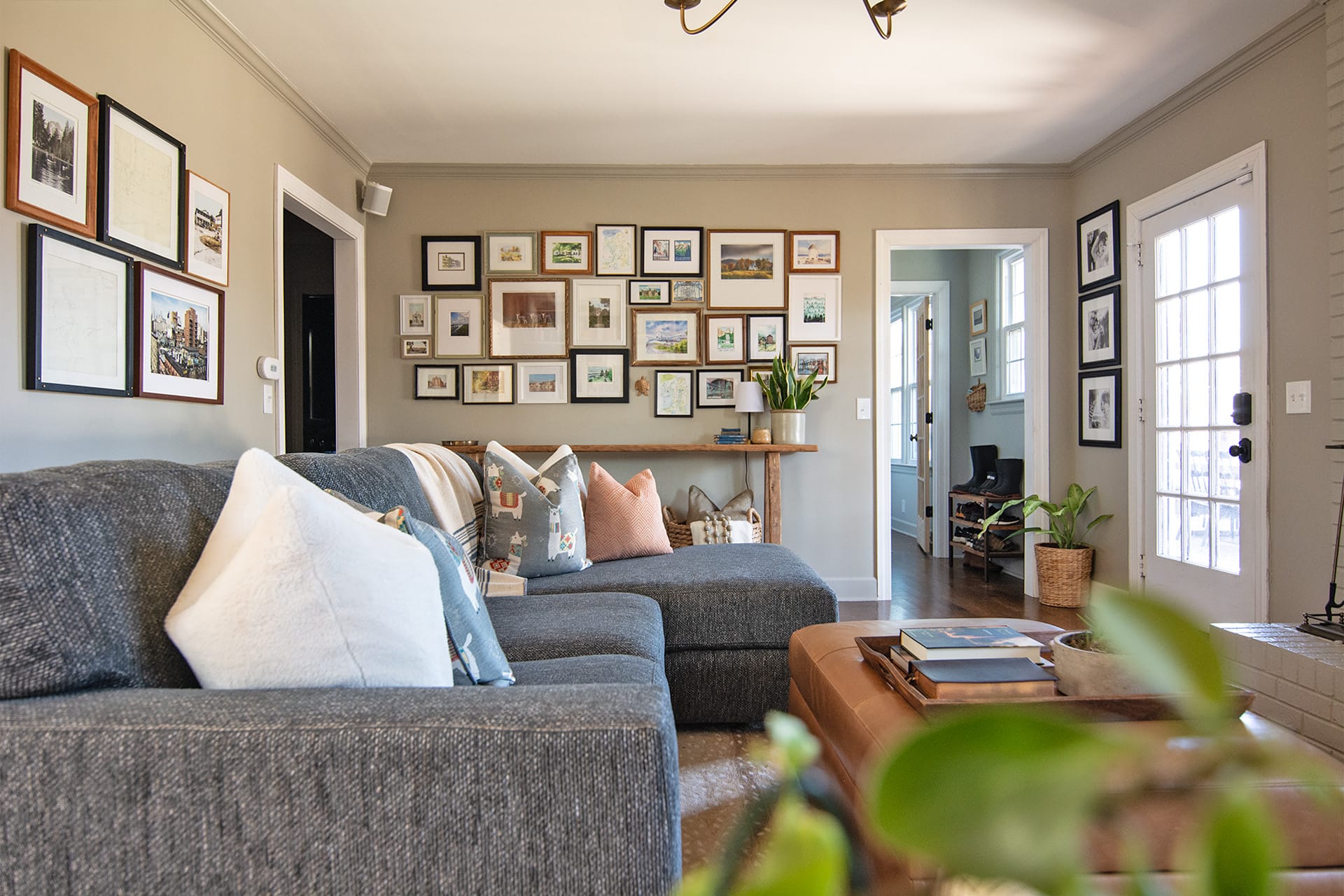 A cozy living room with a gray sectional sofa, colorful throw pillows, a gallery wall with various art pieces, and a hallway leading to other areas of the home.