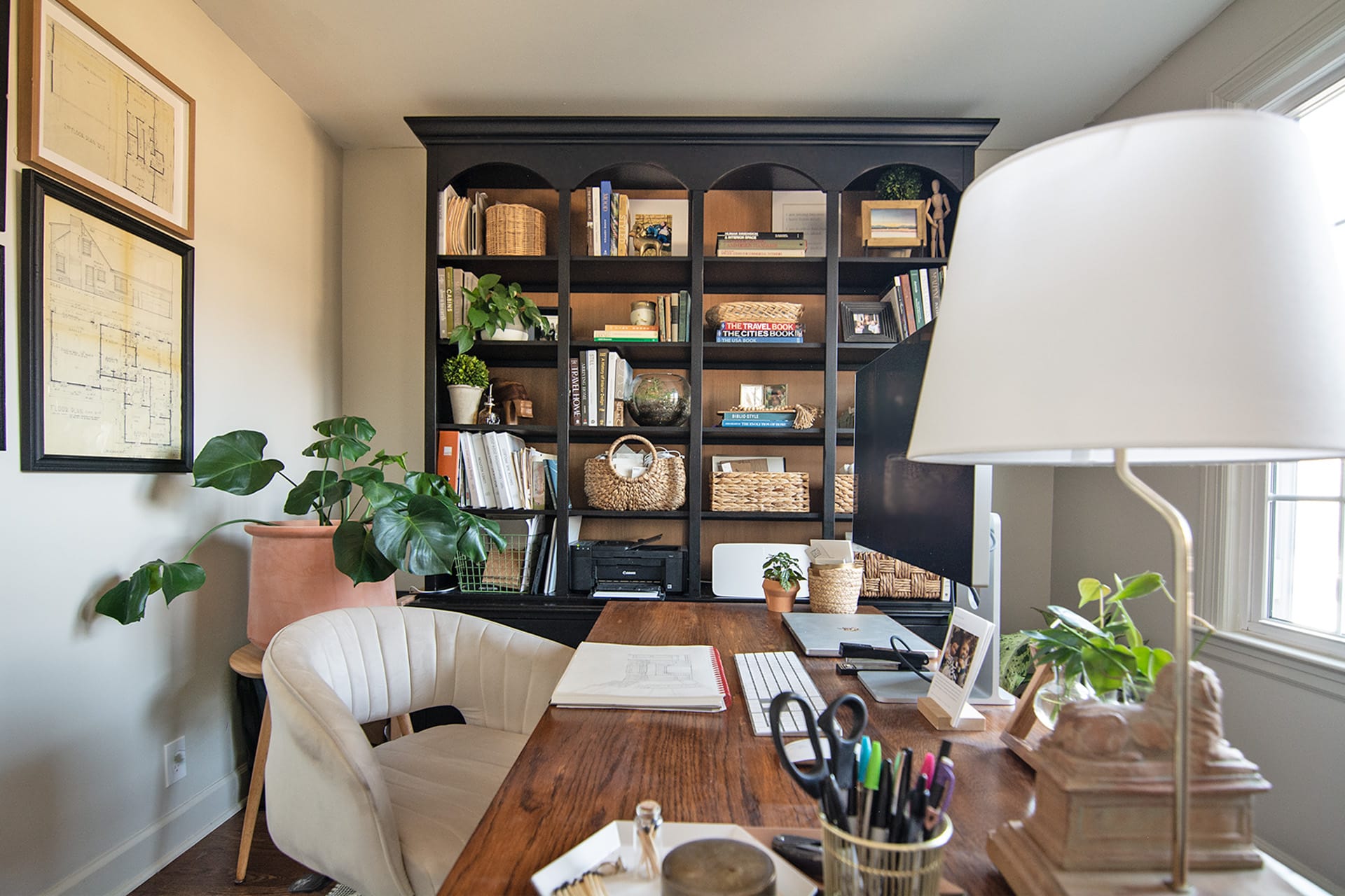 A stylish home office featuring a large wooden desk, a black bookshelf with decor and books, and lush indoor plants for a fresh and inviting workspace.