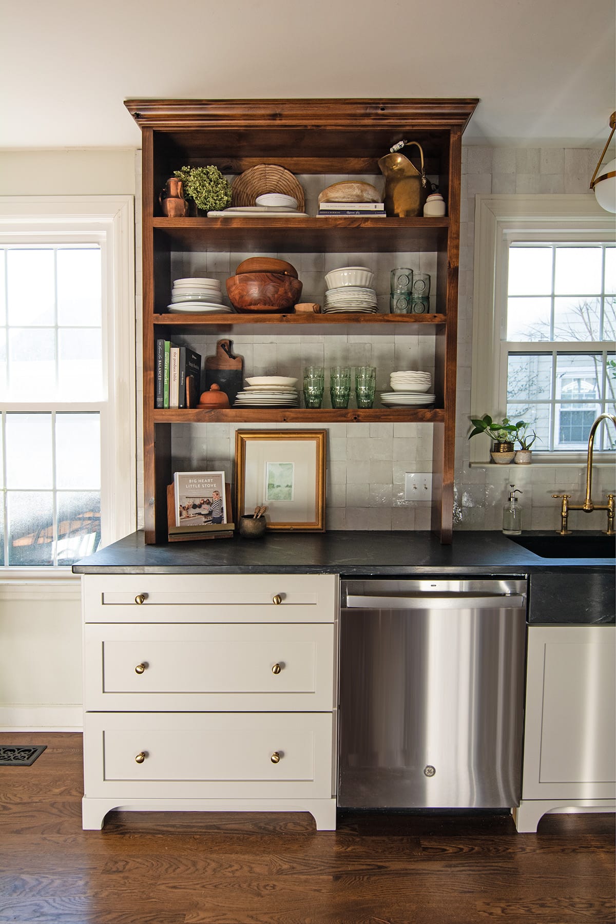 A kitchen with alder open shelving displaying dishes, books, and decor, paired with light cabinetry, brass hardware, and a dark countertop.