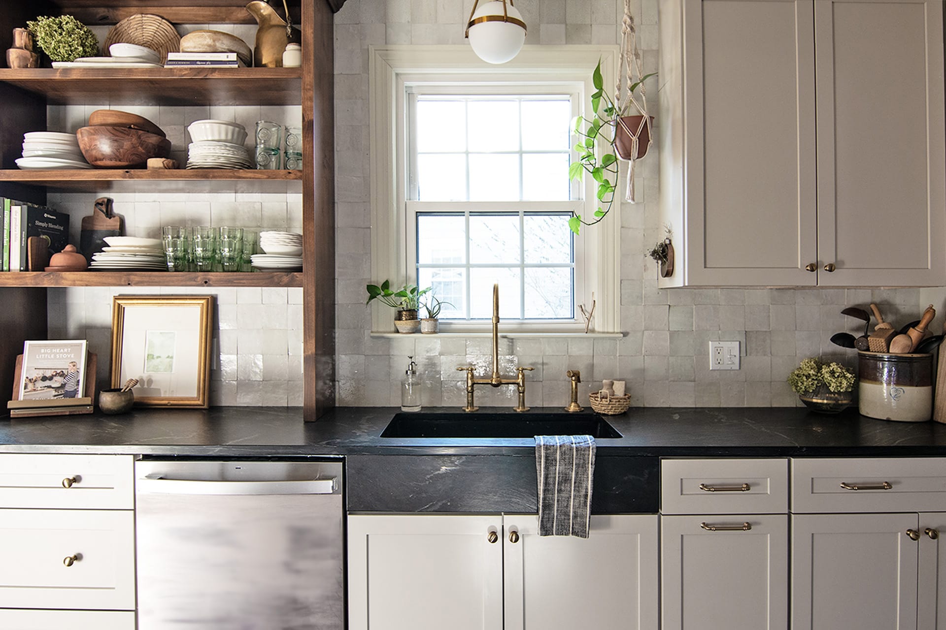 A well-organized kitchen sink wall featuring a brass faucet, black soapstone countertop, open wooden shelves with dishes and decor, and light cream textured tile backsplash.