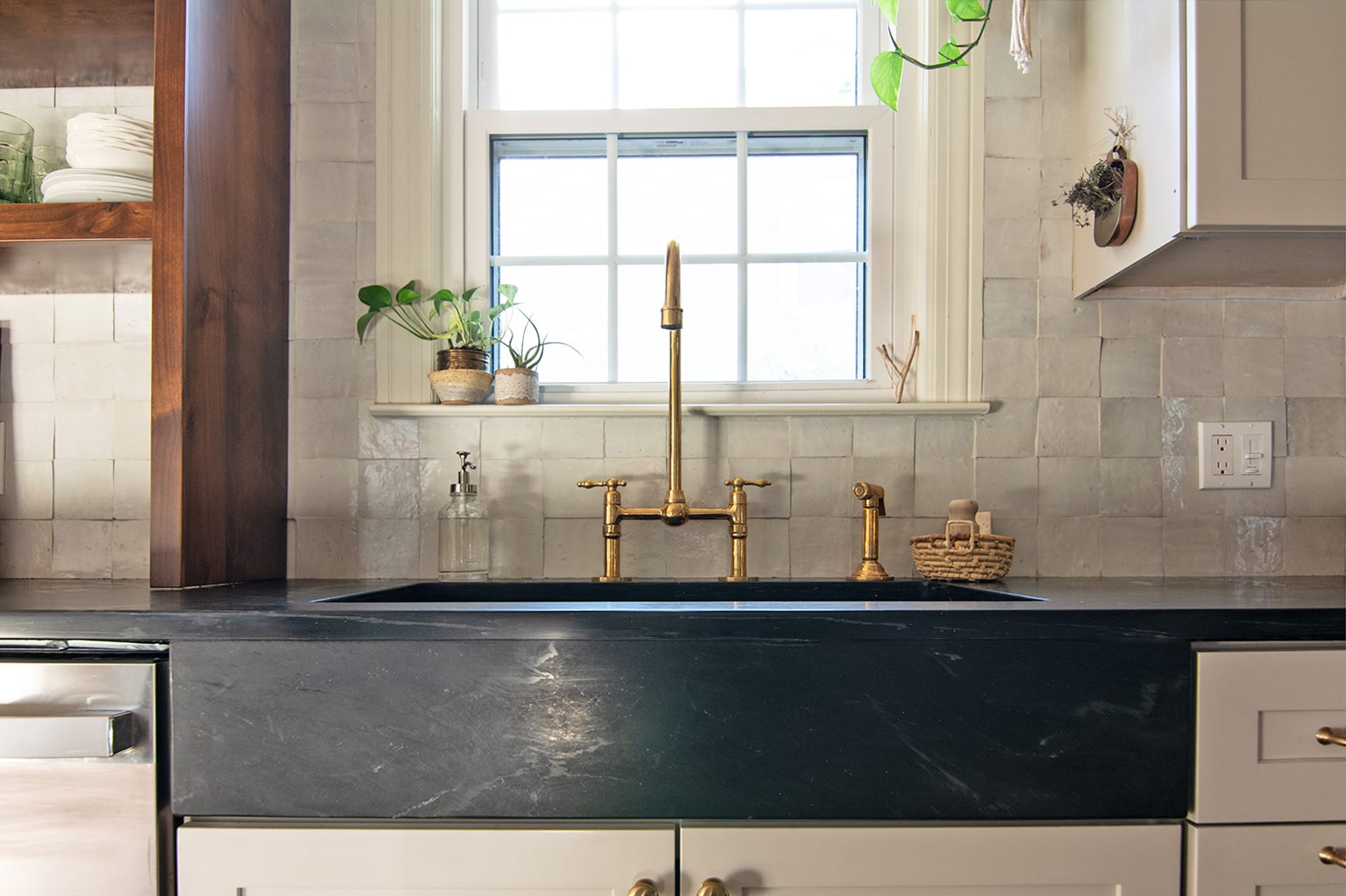 Close-up of a kitchen sink with a brass faucet, black soapstone apron-front sink, and light cream tile backsplash.