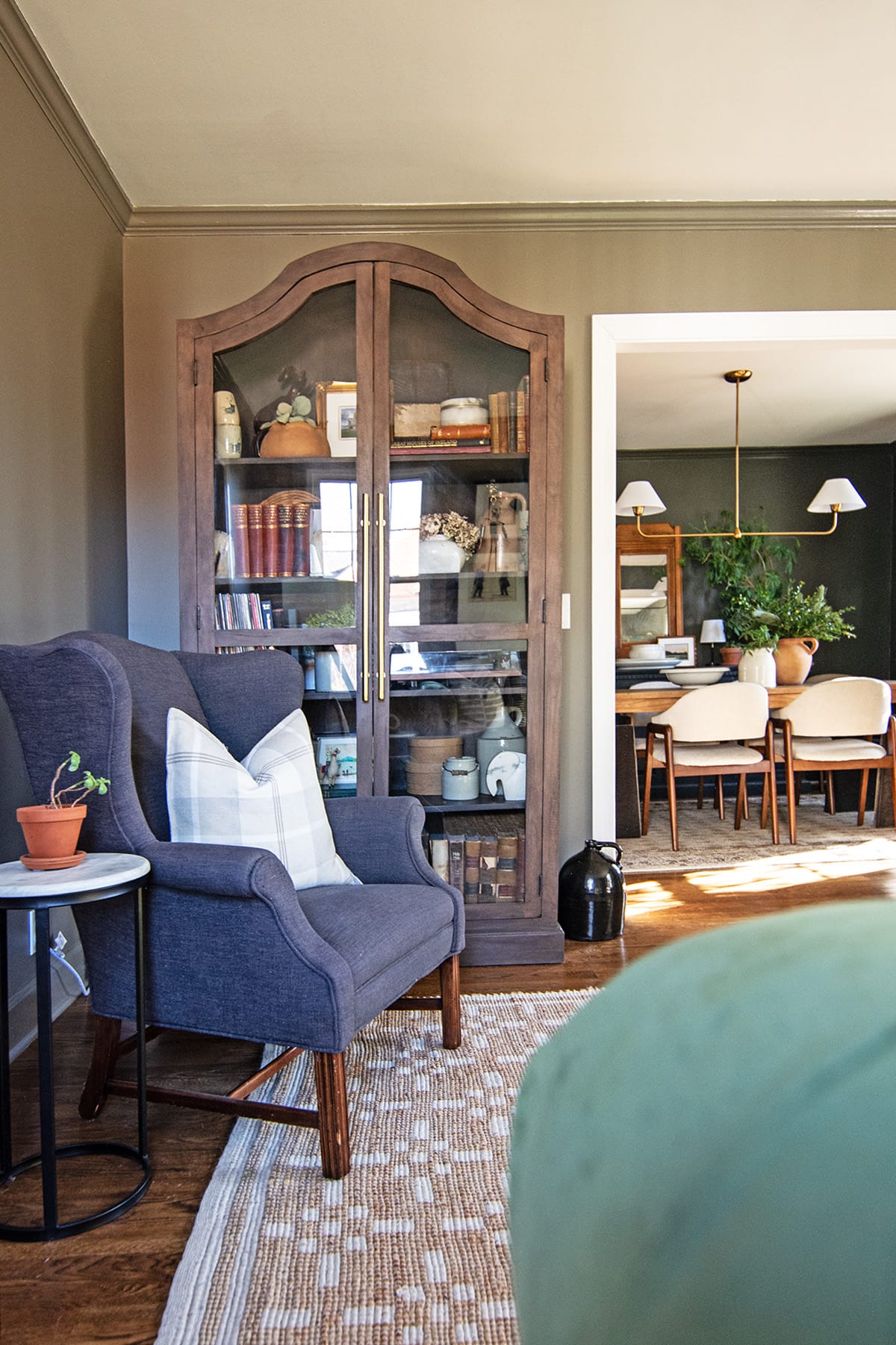 A cozy living room corner featuring a dark wood glass cabinet filled with books and decor, a blue wingback chair with a plaid pillow, and a small marble-top side table.