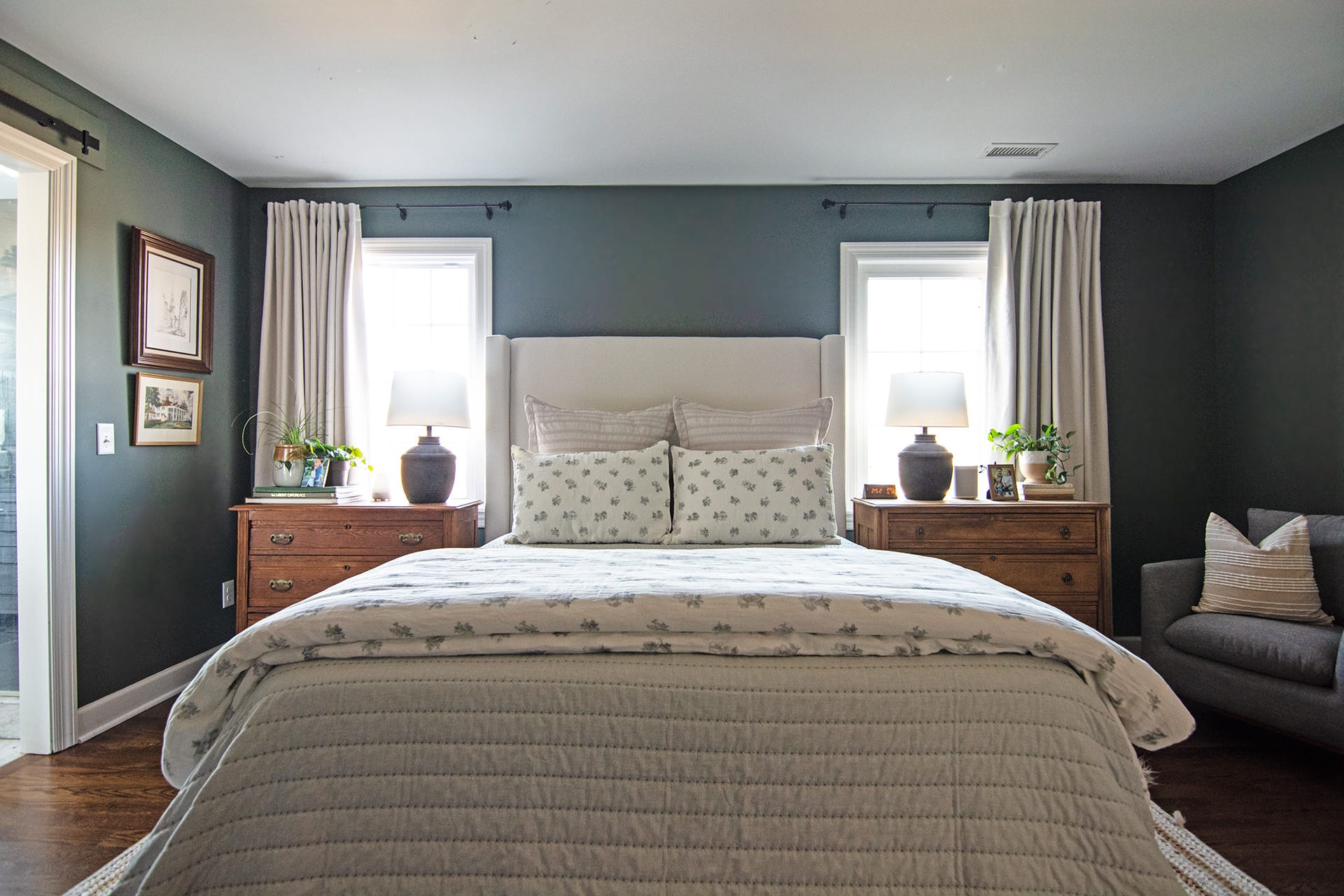 A tranquil primary suite featuring a neutral upholstered bed with floral bedding, wooden nightstands with matching lamps, and soft curtains framing two windows.