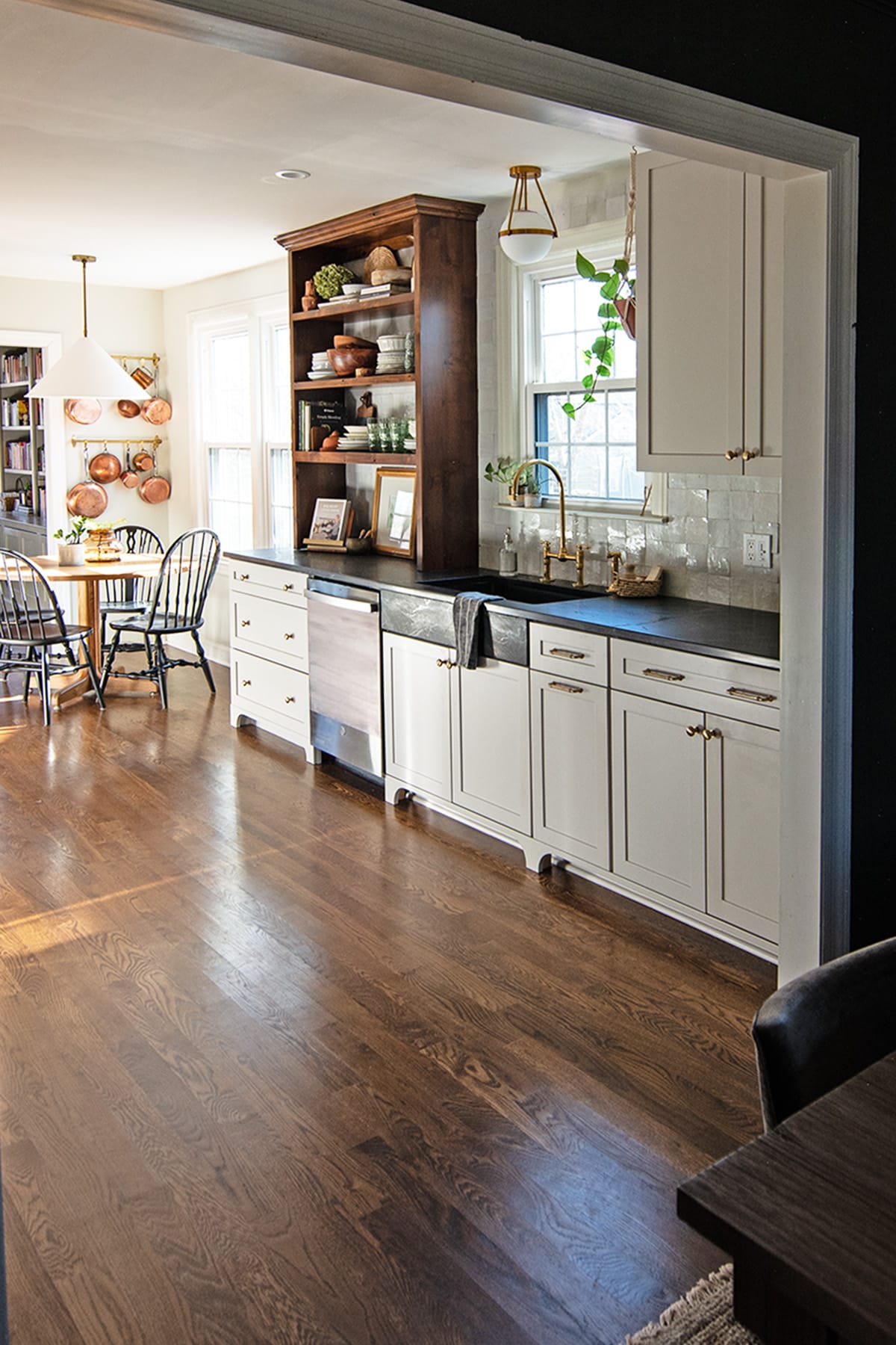 View of a Colonial-style kitchen with light cabinetry, alder wood open shelving, brass hardware, and a farmhouse sink beneath a window, with a dining area visible in the background.