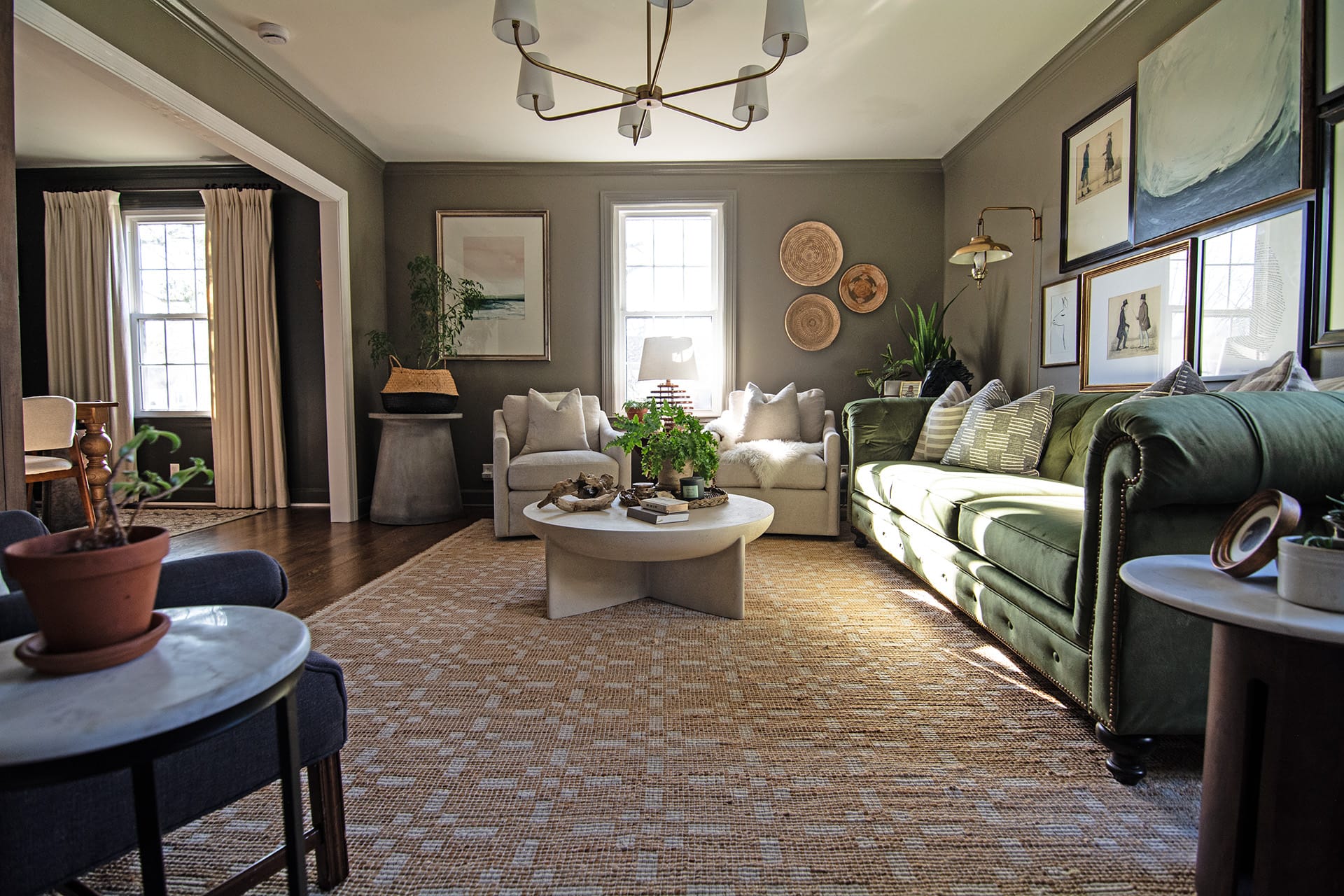 A spacious living room with a green Chesterfield sofa, two neutral armchairs, a round coffee table, and layered decor that combines natural textures and artistic elements.