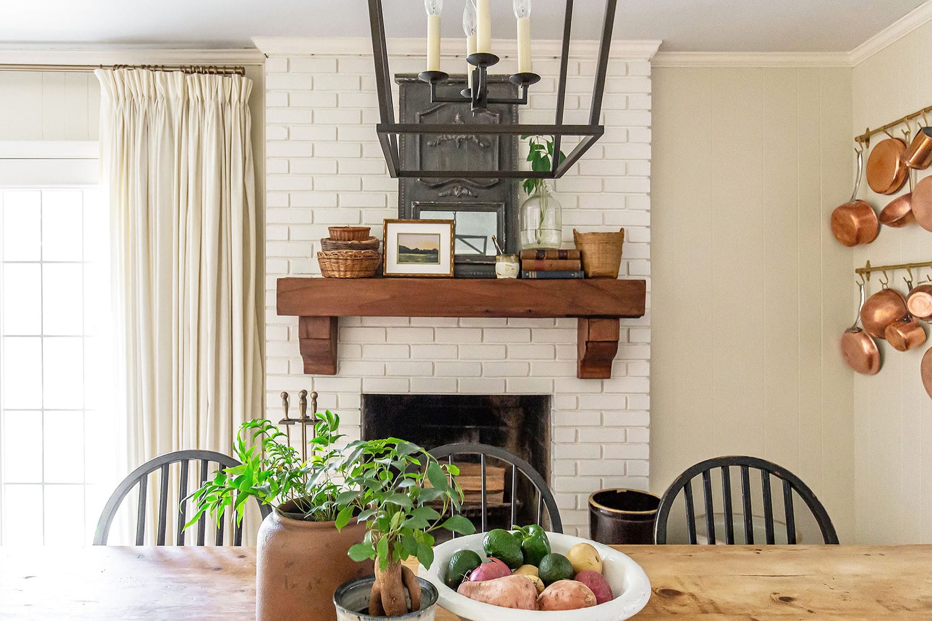 A charming dining room featuring a white brick fireplace with a wooden mantle, adorned with rustic baskets, books, and greenery. A dining table with fresh produce in the foreground complements the warm and inviting space.