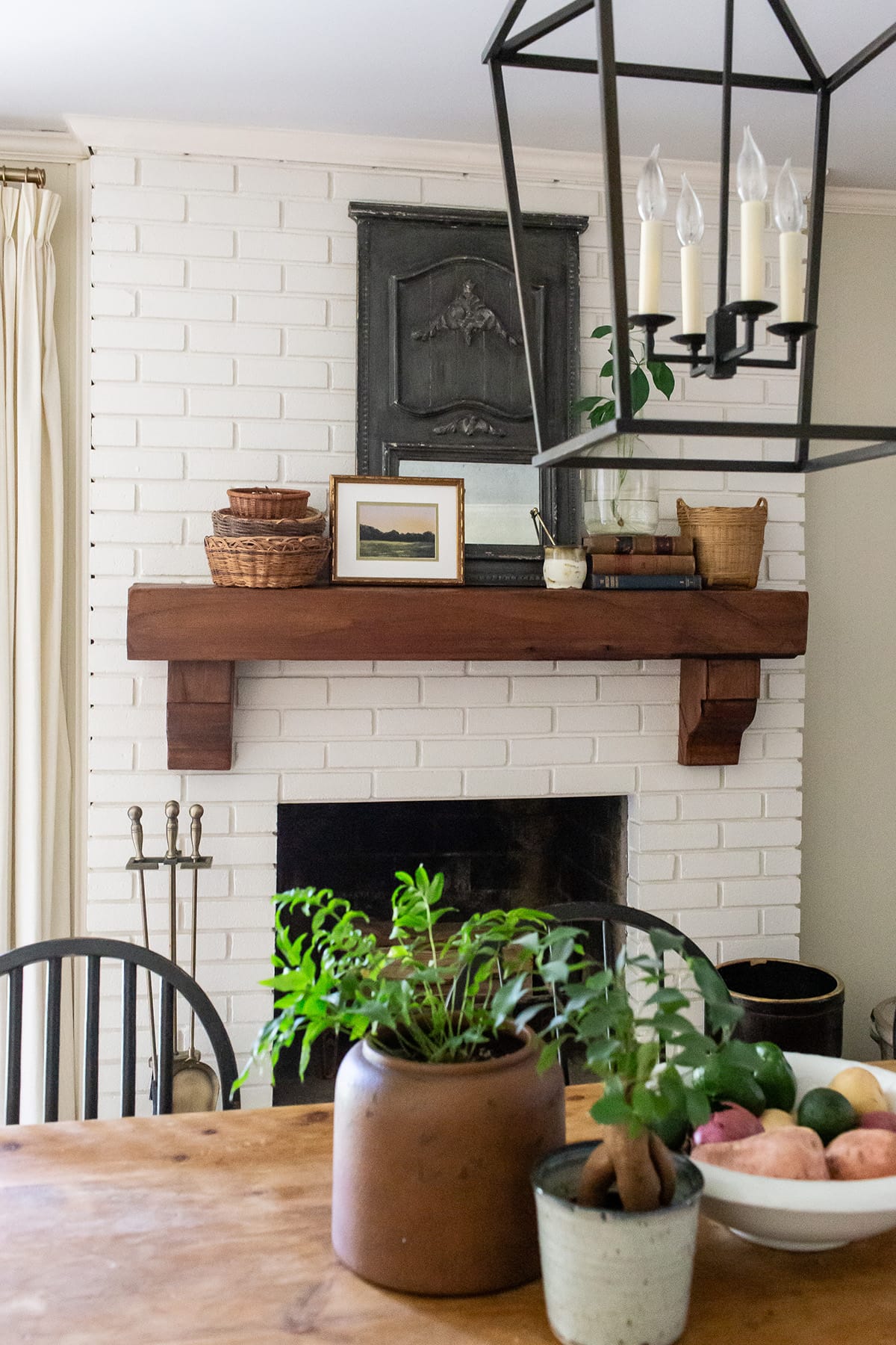 Close-up of a white brick fireplace with a rustic wooden mantle styled with baskets, books, and a black antique panel. Greenery in terracotta and ceramic pots decorates the dining table in the foreground.