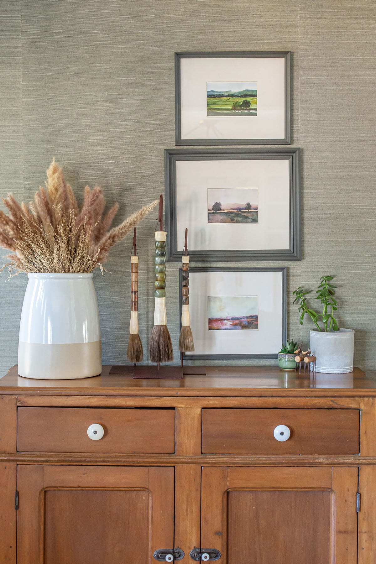 A wooden console table styled with a ceramic vase filled with pampas grass, decorative artisan brushes, framed landscape art, and potted succulents, against a textured green wall.