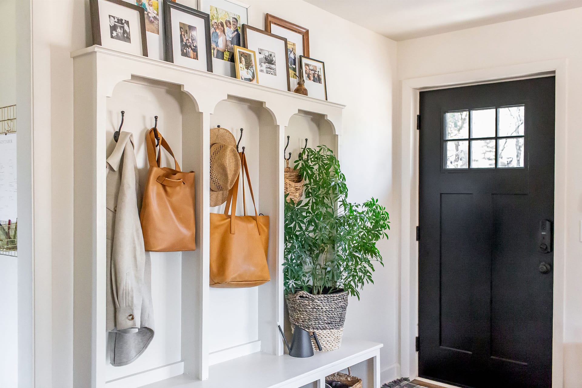 A functional and stylish mudroom entry featuring a white built-in organizer with hooks, baskets, and a bench, accented by a black door and framed family photos.