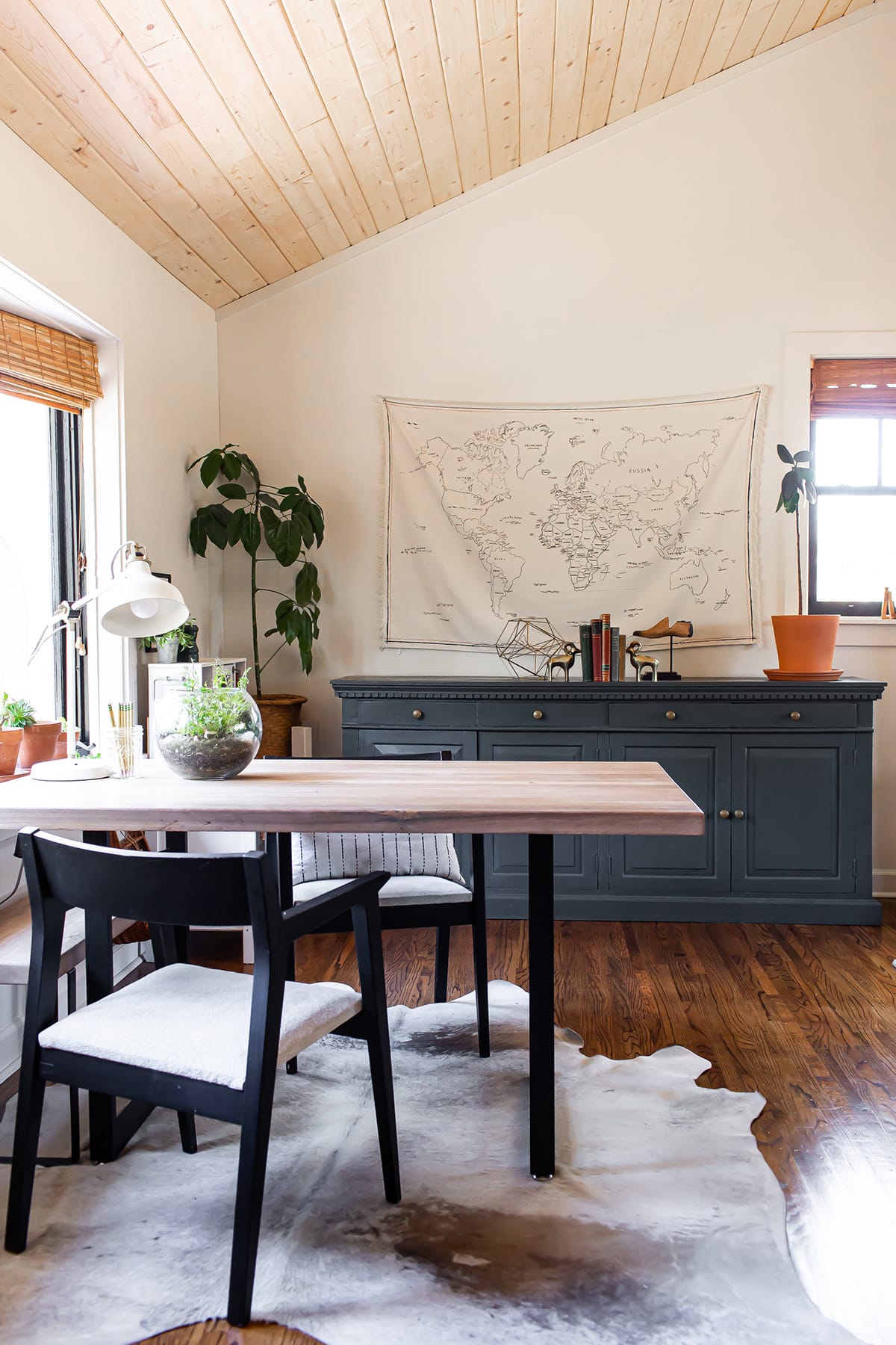 A cozy desk area featuring a wooden table, black modern chairs with white cushions, a green sideboard adorned with decor, and a world map tapestry on the wall.