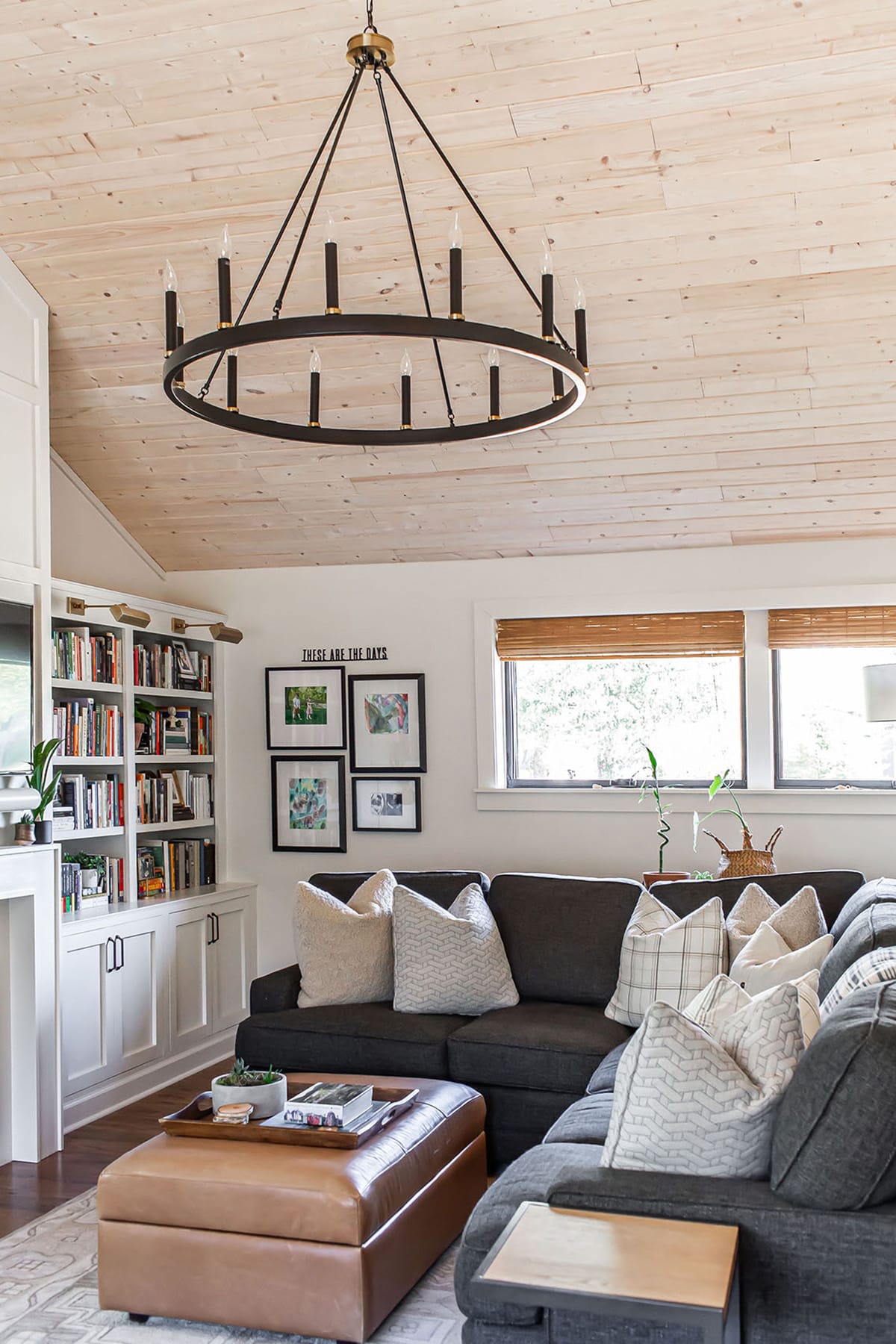 A cozy family room with elevated comfort and rustic elegance featuring a vaulted wood-paneled ceiling, a large circular chandelier, built-in bookshelves, a dark gray sectional sofa, and a leather ottoman.