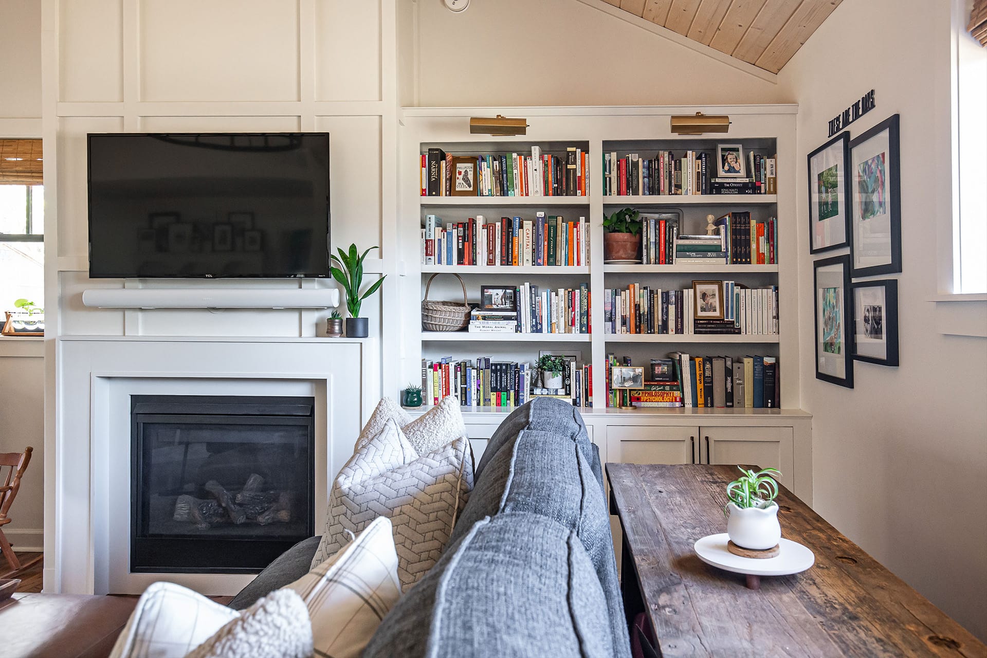 A cozy family room featuring a white fireplace with a mounted TV, built-in bookshelves filled with books and decor, and a gray sectional sofa with decorative pillows.