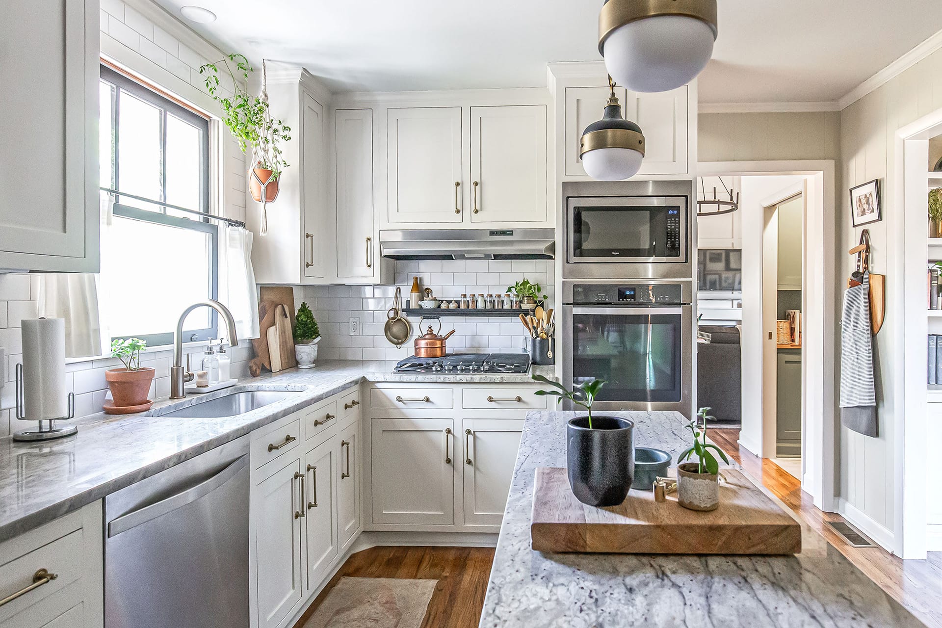 Bright and inviting kitchen featuring marble countertops, a gas stovetop with a stainless steel hood, white cabinetry with brass hardware, and a hanging plant by the window.