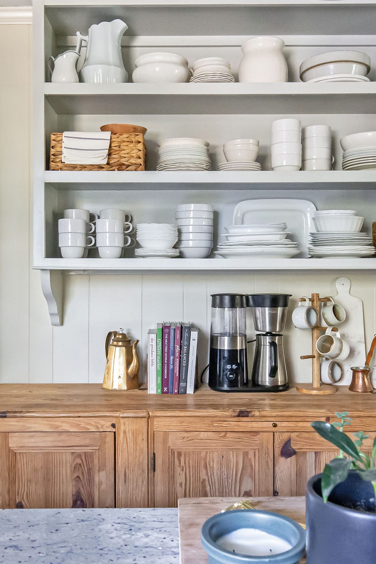 Open shelving with neatly arranged white dinnerware, a brass coffee pot, books, and a modern coffee maker set on a wooden cabinet.