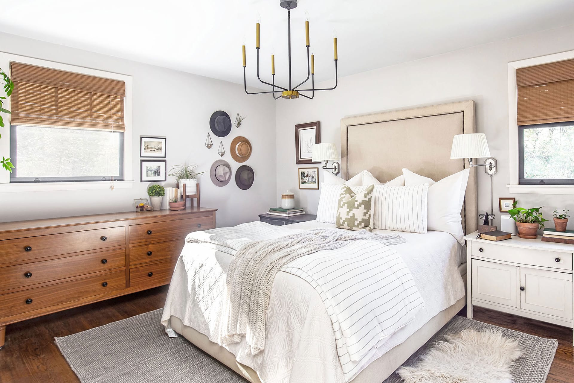 A serene primary bedroom with a neutral upholstered bed, layered white bedding, and cozy textiles, complemented by a wooden dresser, gallery wall with hats and framed art, and potted greenery.