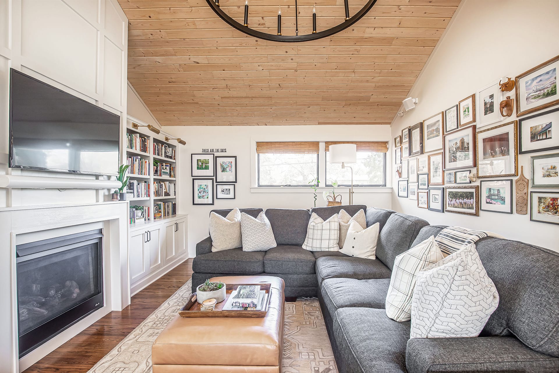 A cozy family room with a vaulted wood ceiling, a sectional sofa, a gallery wall, a built-in bookcase, and a white fireplace topped with a mounted TV.