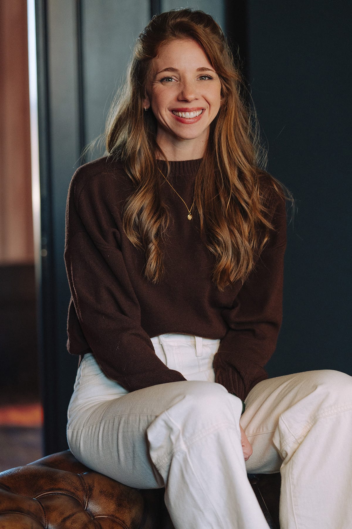 Portrait of Kaylee Pauley with long, wavy hair wearing a dark brown sweater and white pants, sitting on a tufted leather seat against a dark background.
