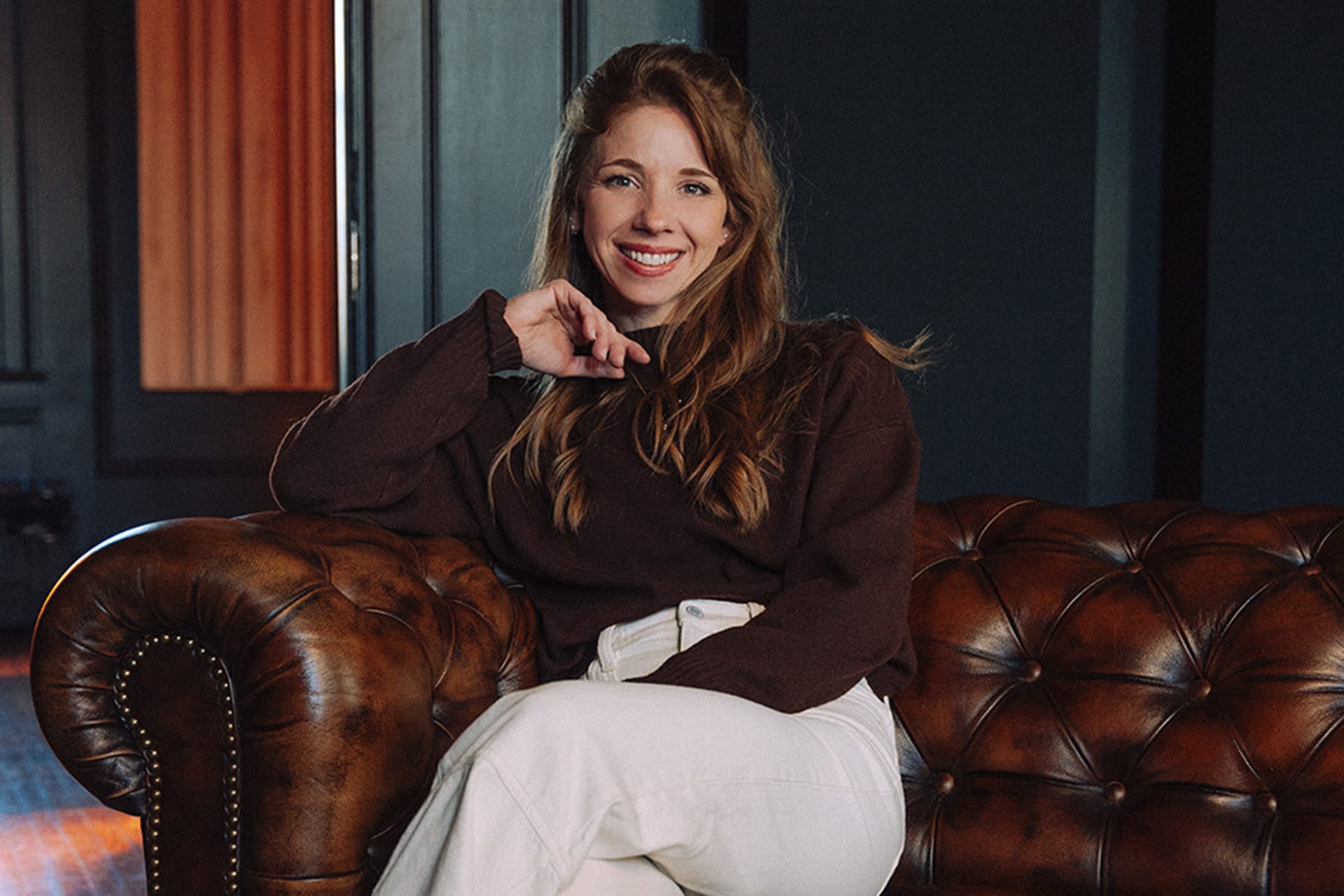 Kaylee Pauley seated on a tufted leather couch, smiling warmly in a stylish and cozy interior setting.